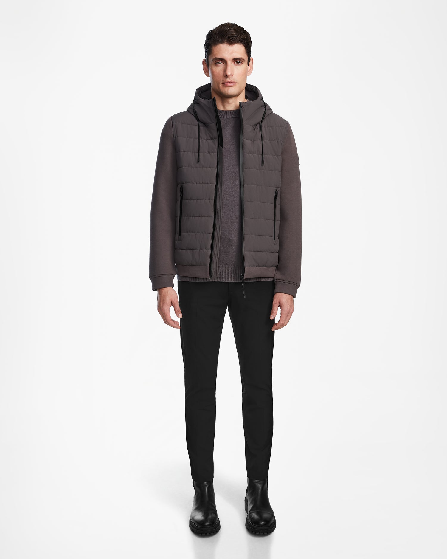 A man faces forward wearing the Rudsak ELIAS - M124002 Grey men’s quilted jacket with a hood, paired with a brownish-gray sweater, black pants, and black boots against a plain white background.