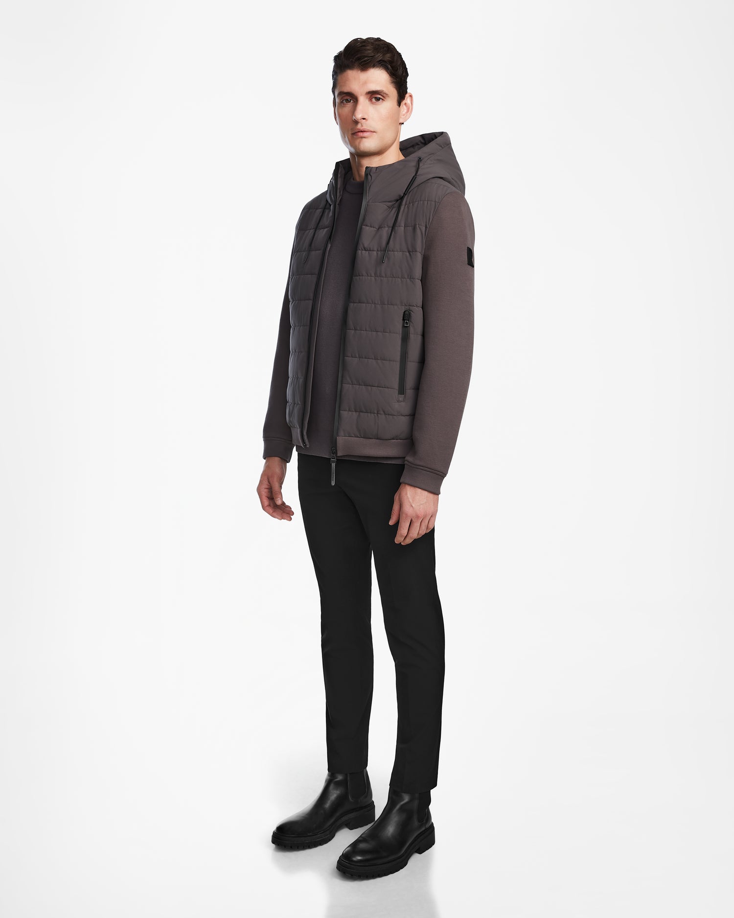 A man stands against a white background wearing the Rudsak ELIAS - M124002 Grey quilted jacket over a long-sleeve shirt, slim black pants, and black boots, looking straight ahead with a neutral expression.