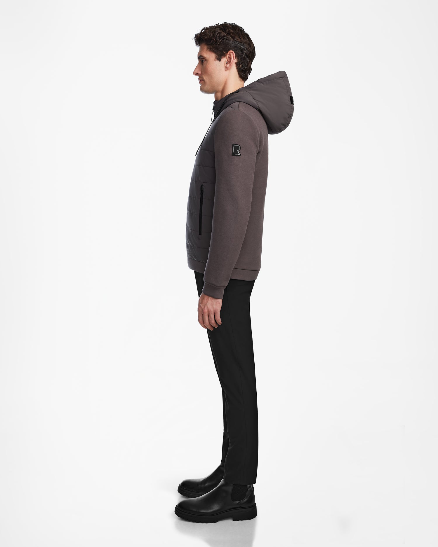 A man stands in profile against a white background, wearing Rudsaks ELIAS - M124002 Grey quilted jacket, paired with black pants and black boots.