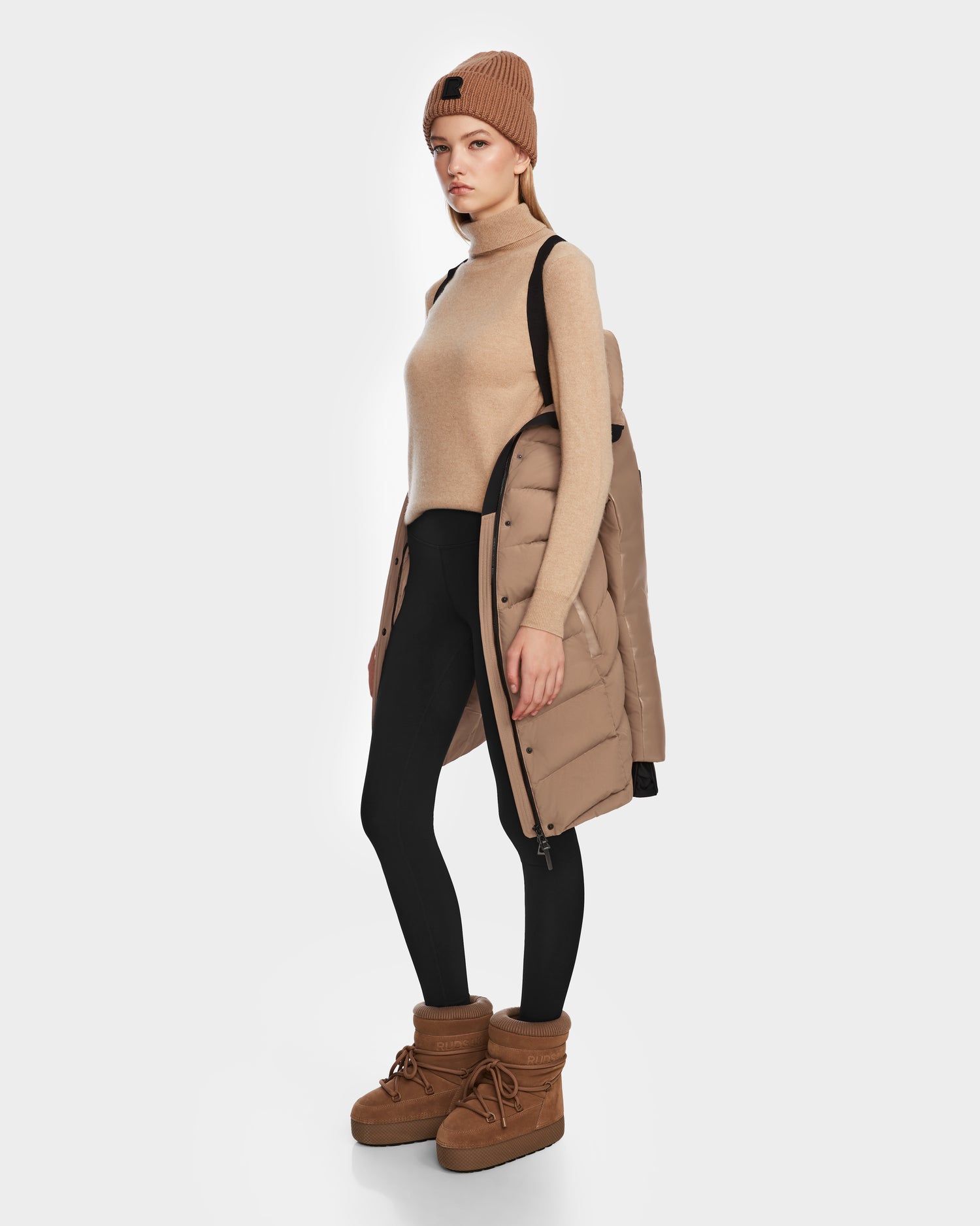 A woman in a tan beanie, sleeveless turtleneck sweater, black leggings, and Rudsak ELSA - L123535 Sand RDS-certified down puffer coat draped over her shoulders stands in tan winter boots against a white background.
