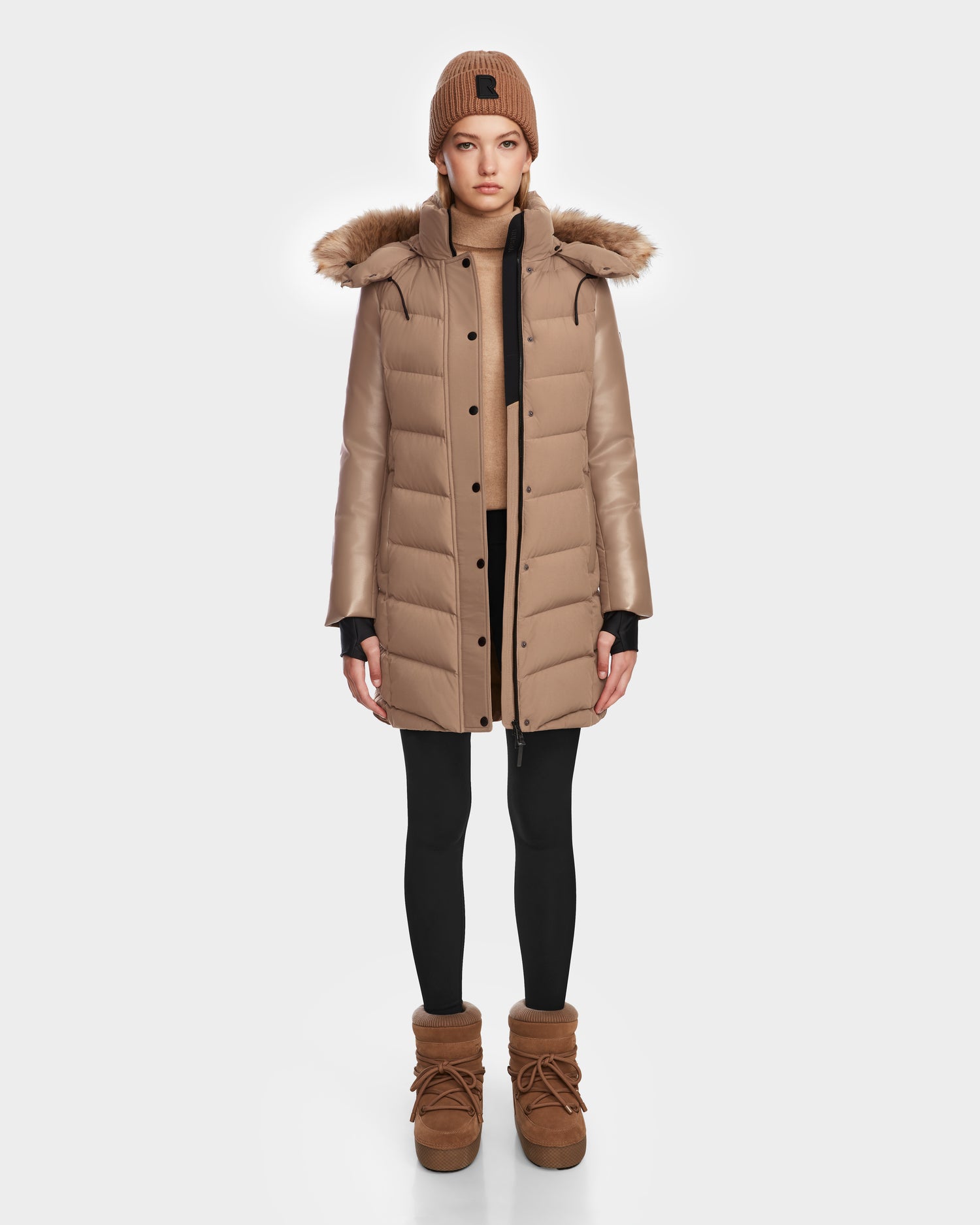 A person stands facing forward, wearing the Rudsak ELSA - L123535 Sand women’s quilted down puffer coat with fur-trimmed hood, a matching knit beanie, tan sweater, black leggings, and tan boots against a plain white background.