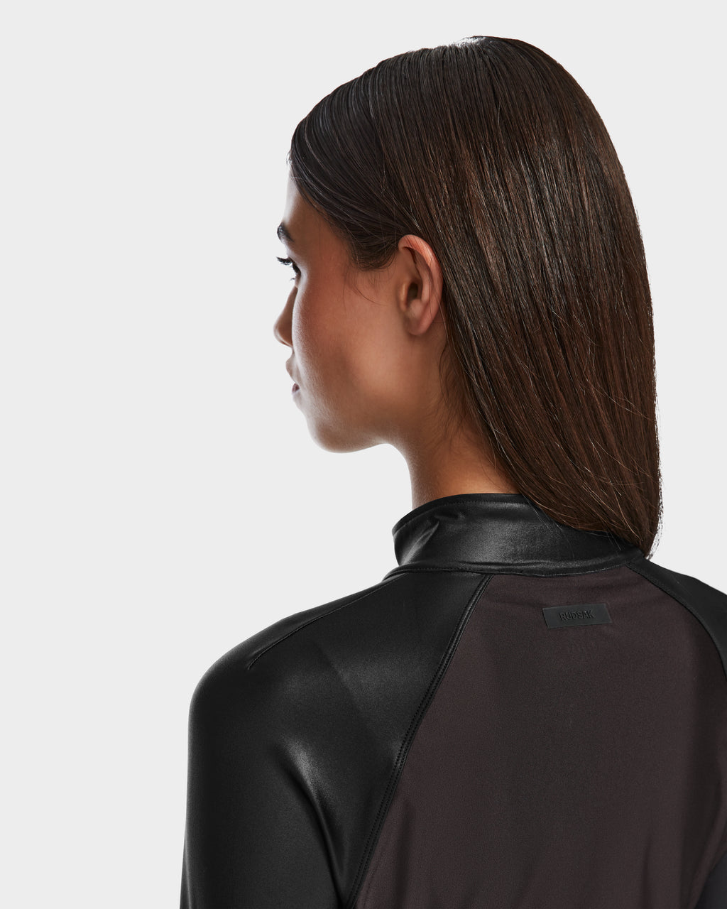 A woman with straight brown hair is shown from the back and side wearing the Rudsak EVA - 8524514 Black jacket, featuring shiny stretch fabric, a raised collar, and waterproof zipper against a plain white background.
