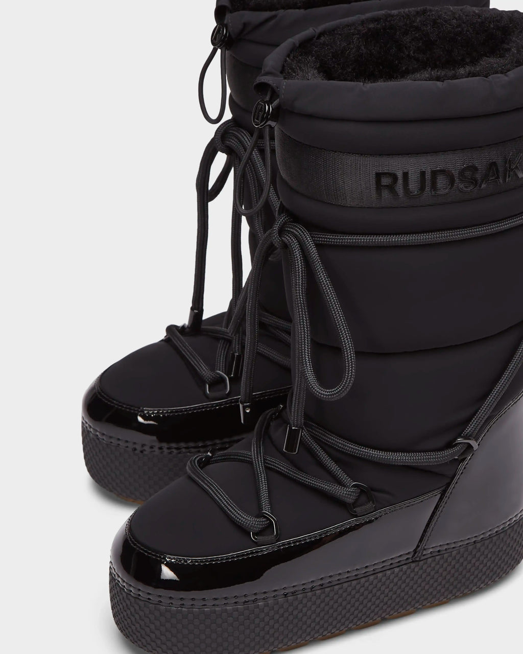 Close-up of Rudsaks FARAH L - 8224542 Black womens winter boots featuring shiny, textured soles, lace-up fronts, plush inner lining, and Rudsak branding near the top of the shaft.