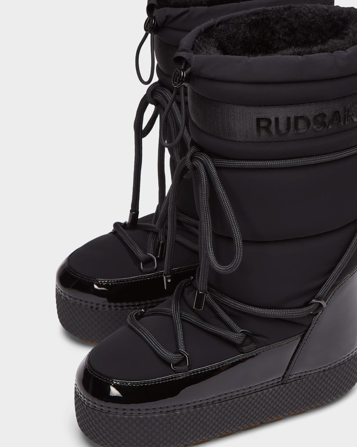 Close-up of Rudsaks FARAH L - 8224542 Black womens winter boots featuring shiny, textured soles, lace-up fronts, plush inner lining, and Rudsak branding near the top of the shaft.