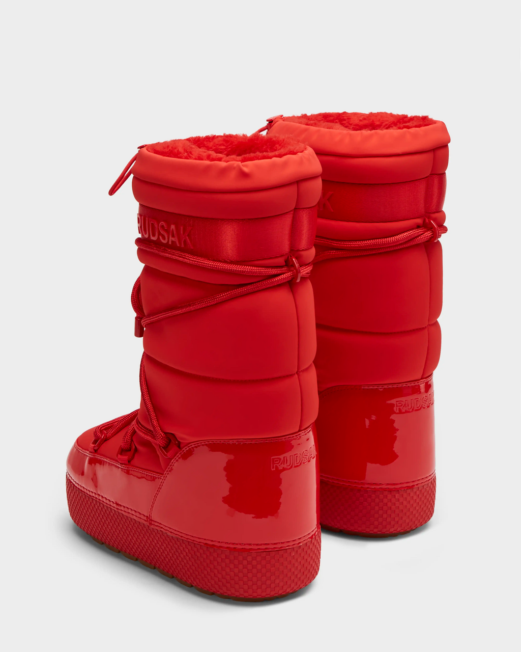 The FARAH - 8223501 Flame Red by Rudsak are bright red, high-top winter boots with puffy, quilted sides, lace-up fronts, glossy rubber soles, faux fur lining for warmth, and Rudsak embossed near the tops and heels.