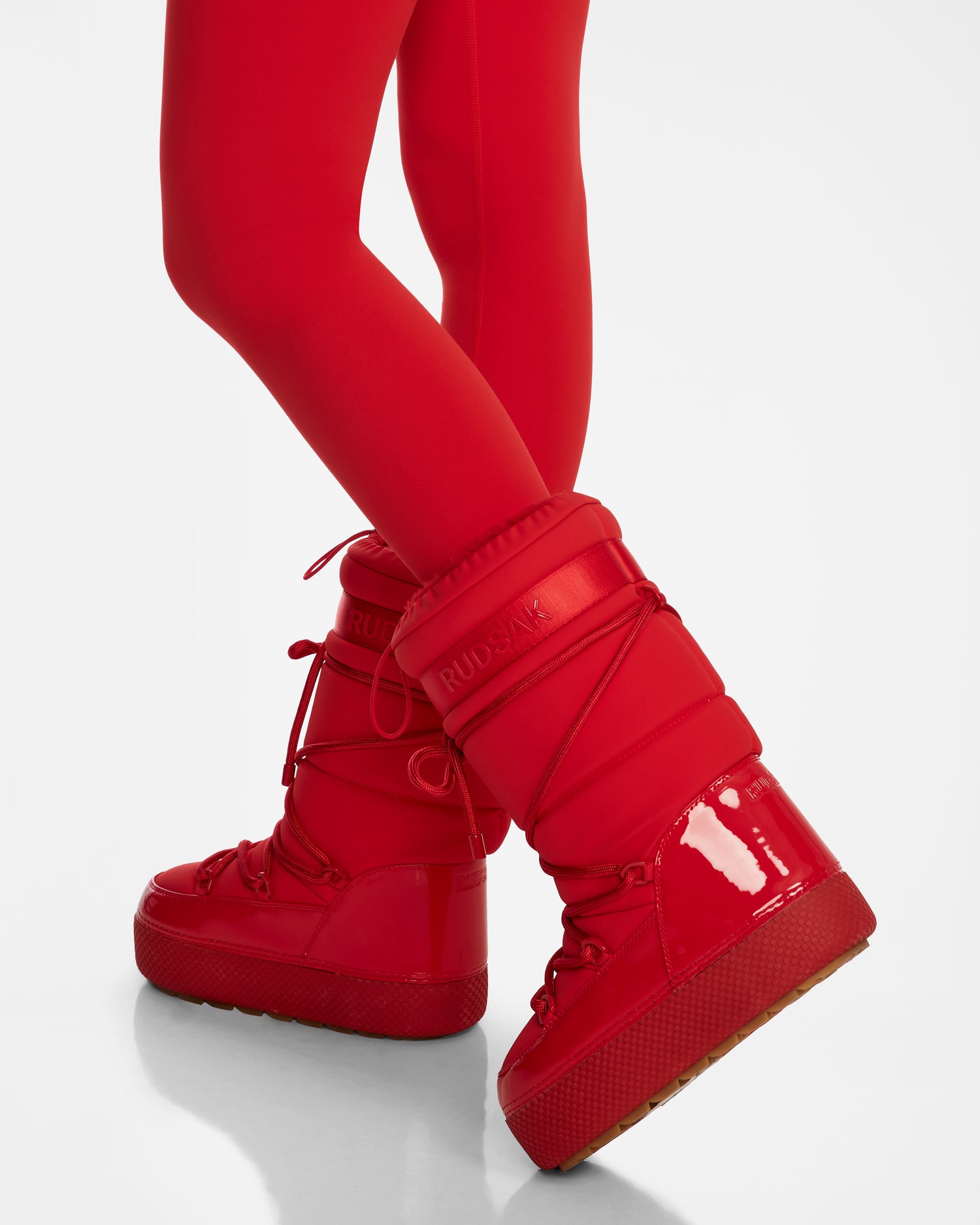 A person models the Rudsak FARAH - 8223501 Flame Red tall, shiny moon boots with faux fur lining and matching red leggings, standing cross-legged against a plain white background.