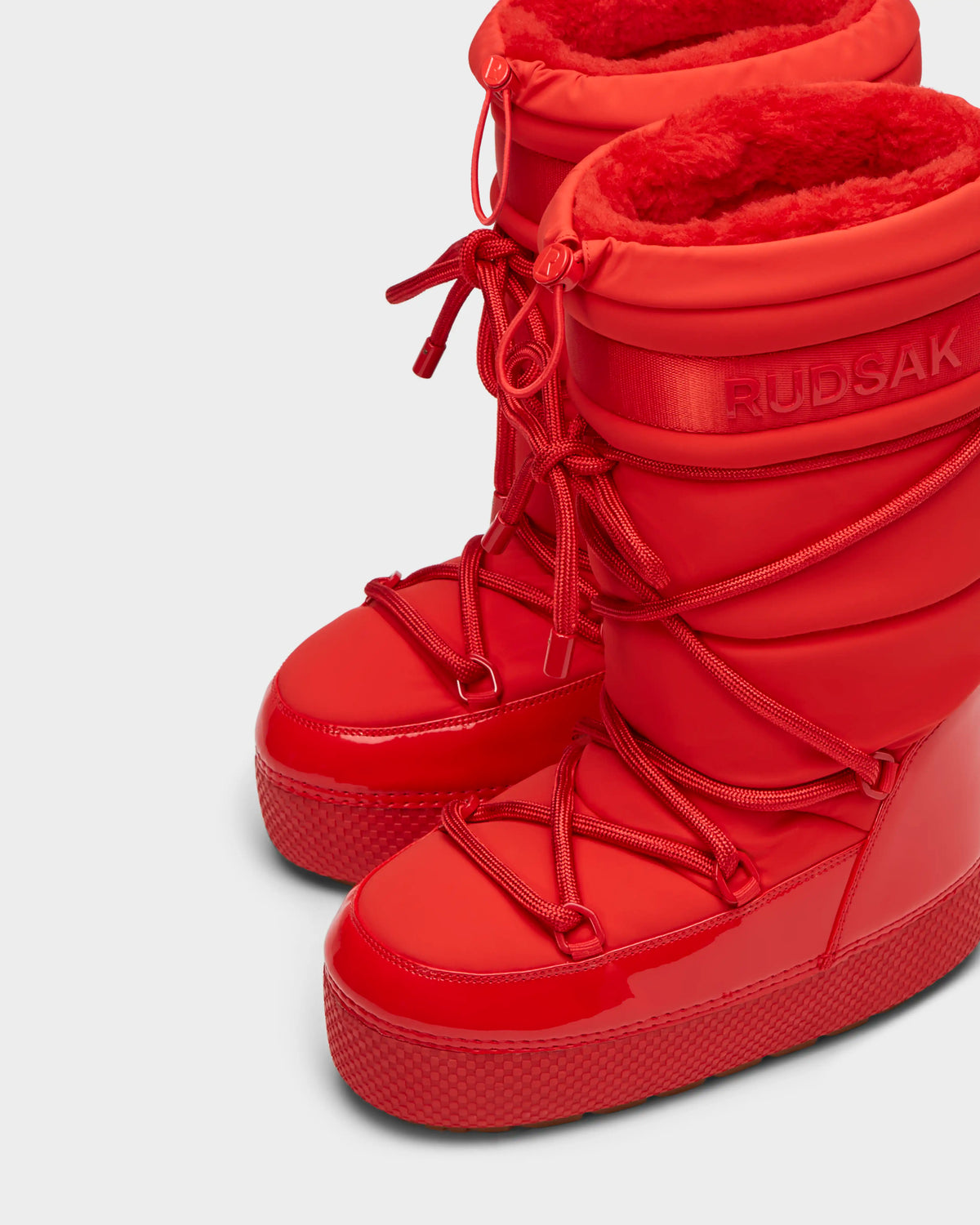 The FARAH - 8223501 Flame Red boots by Rudsak are tall, bright red moon boots with glossy, textured soles, thick red laces, and plush faux fur lining. RUDSAK branding near the top adds style and comfort.