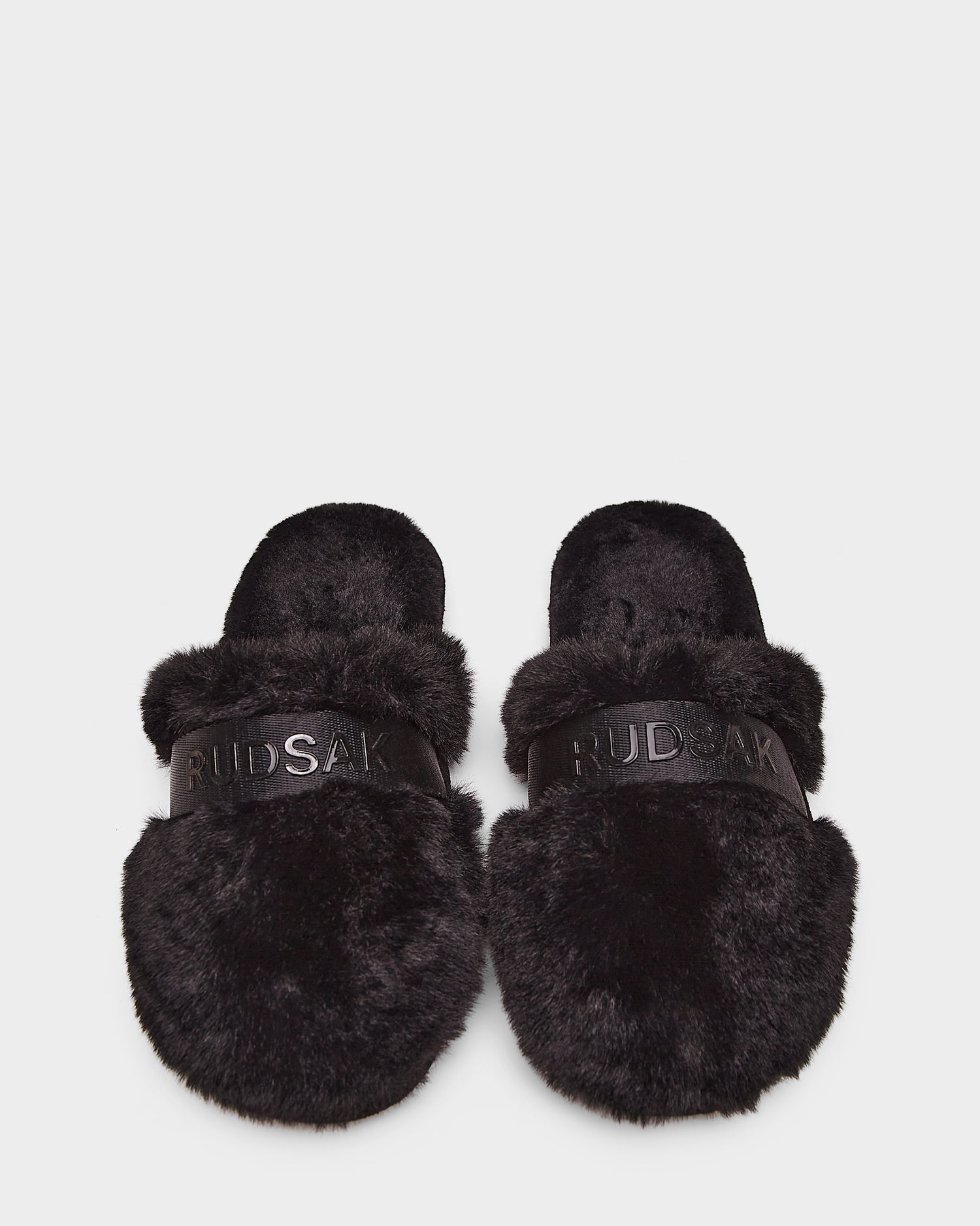 The Rudsak FLUFF - L223509 Black slippers feature cozy black faux-fur and RUDSAK branding across the straps, shown on a white background.