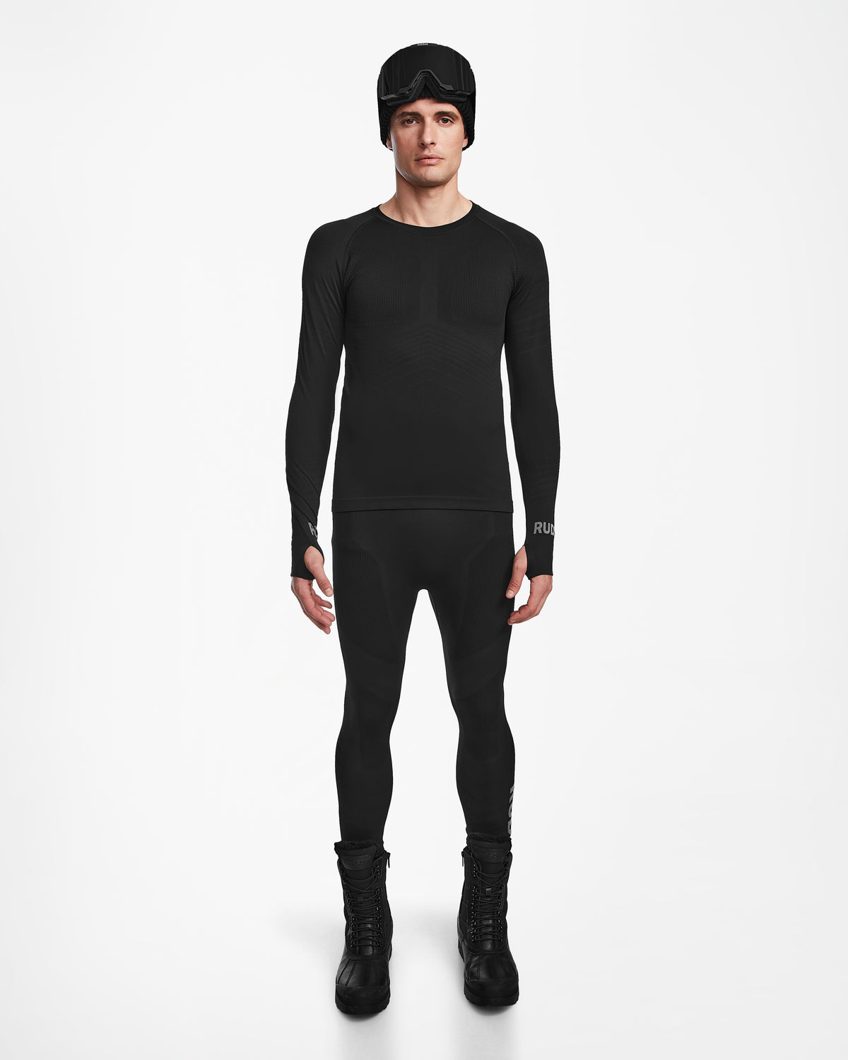 A man stands facing forward wearing the Rudsak FRANC - 6524541 Black fitted men’s thermal set with matching leggings, tall boots, and a black beanie with goggles, posed against a plain white background.