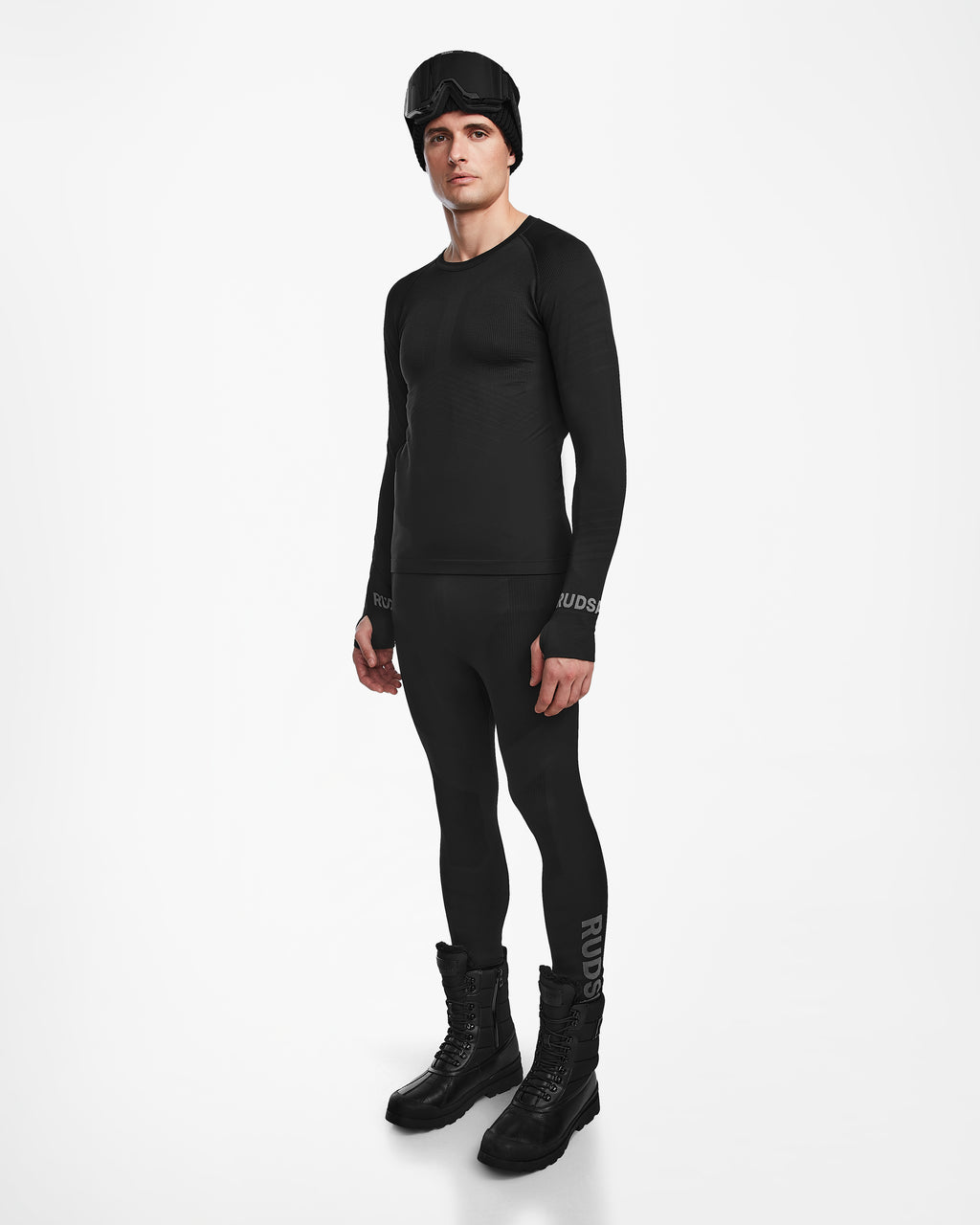 A man wears the Rudsak FRANC - 6524541 Black men’s thermal set—black stretch base layer top and bottom—paired with black snow boots and a black beanie with ski goggles, standing against a plain white background.