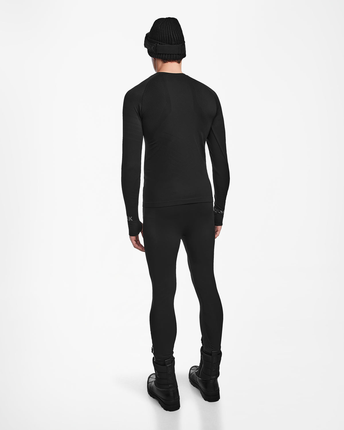 A person showcases the back view of the Rudsak FRANC - 6524541 Black ski underlayer outfit against a plain white background, wearing a black beanie, long-sleeve top, leggings, and boots.