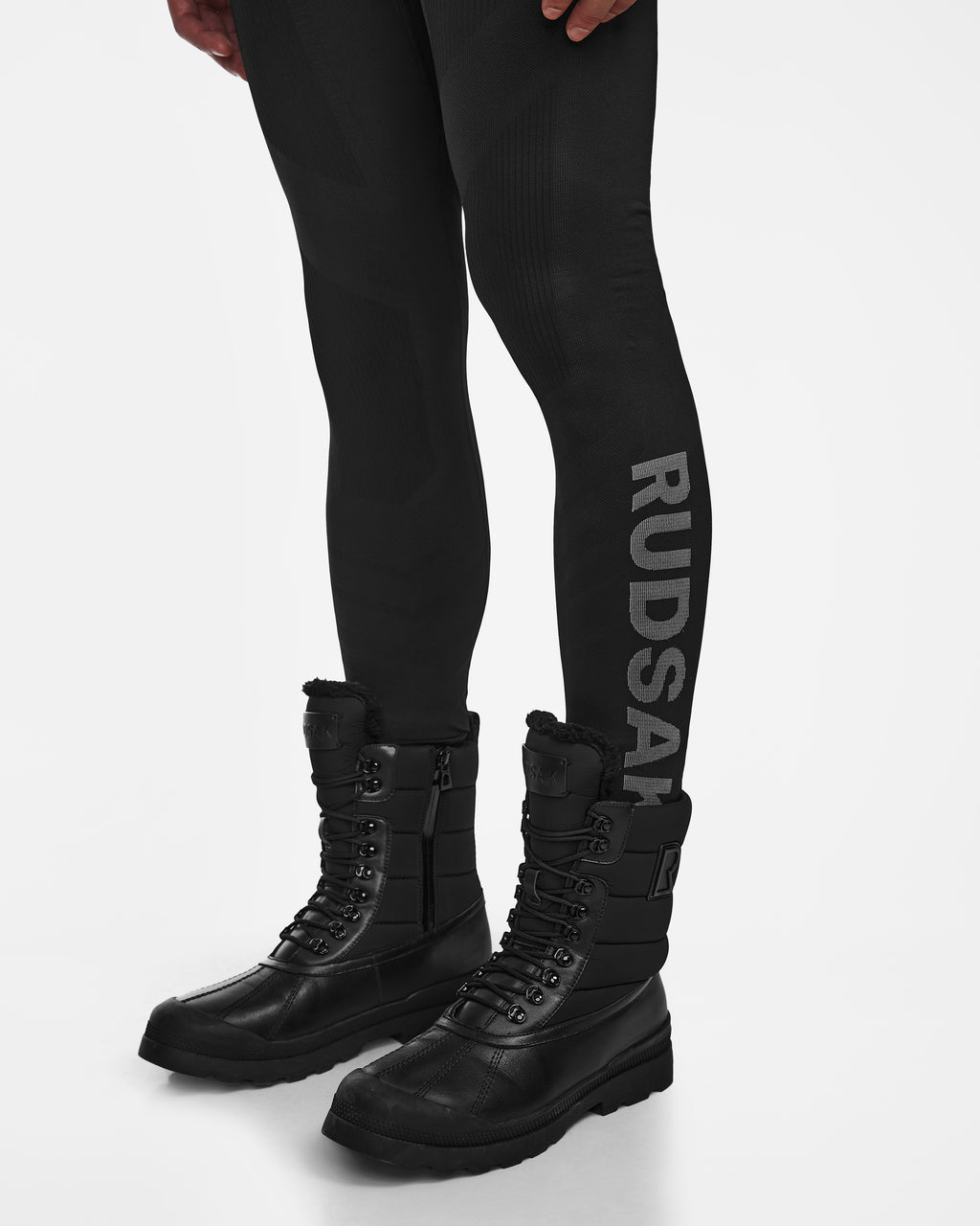 A person wearing Rudsak FRANC - 6524541 black stretch base layer leggings with RUDSAK in gray on one leg and black lace-up winter boots, standing on a white background.