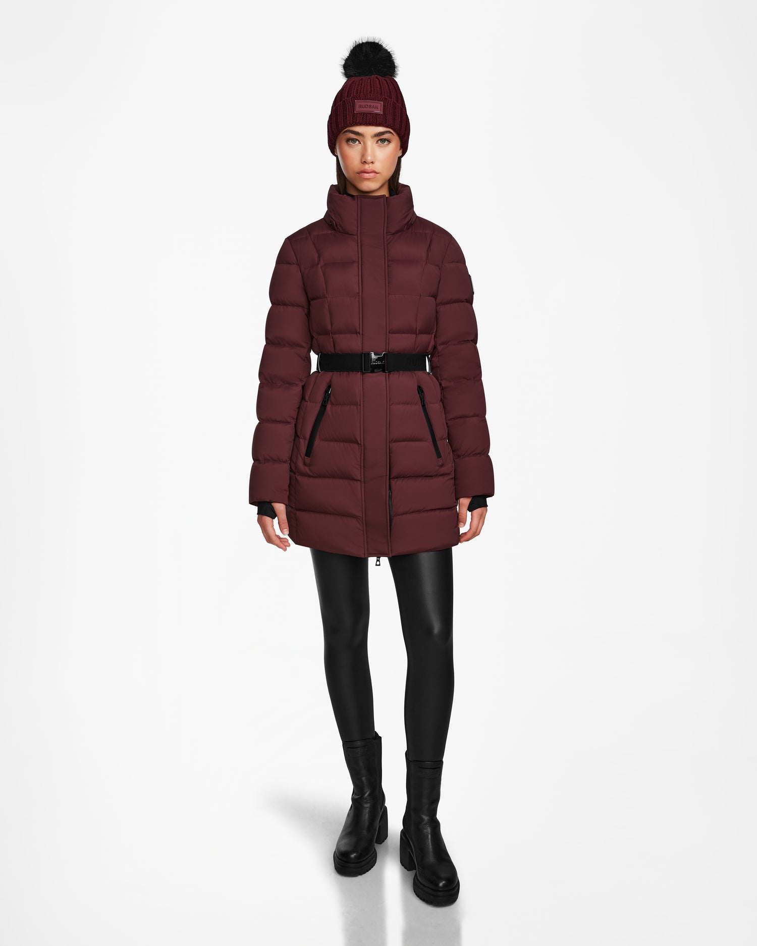 A woman models the Rudsak FRIDA M - L124524 Wine, a stylish womens mid-length down jacket with a removable hood, black belt, and matching beanie, paired with black leggings and boots against a white background.