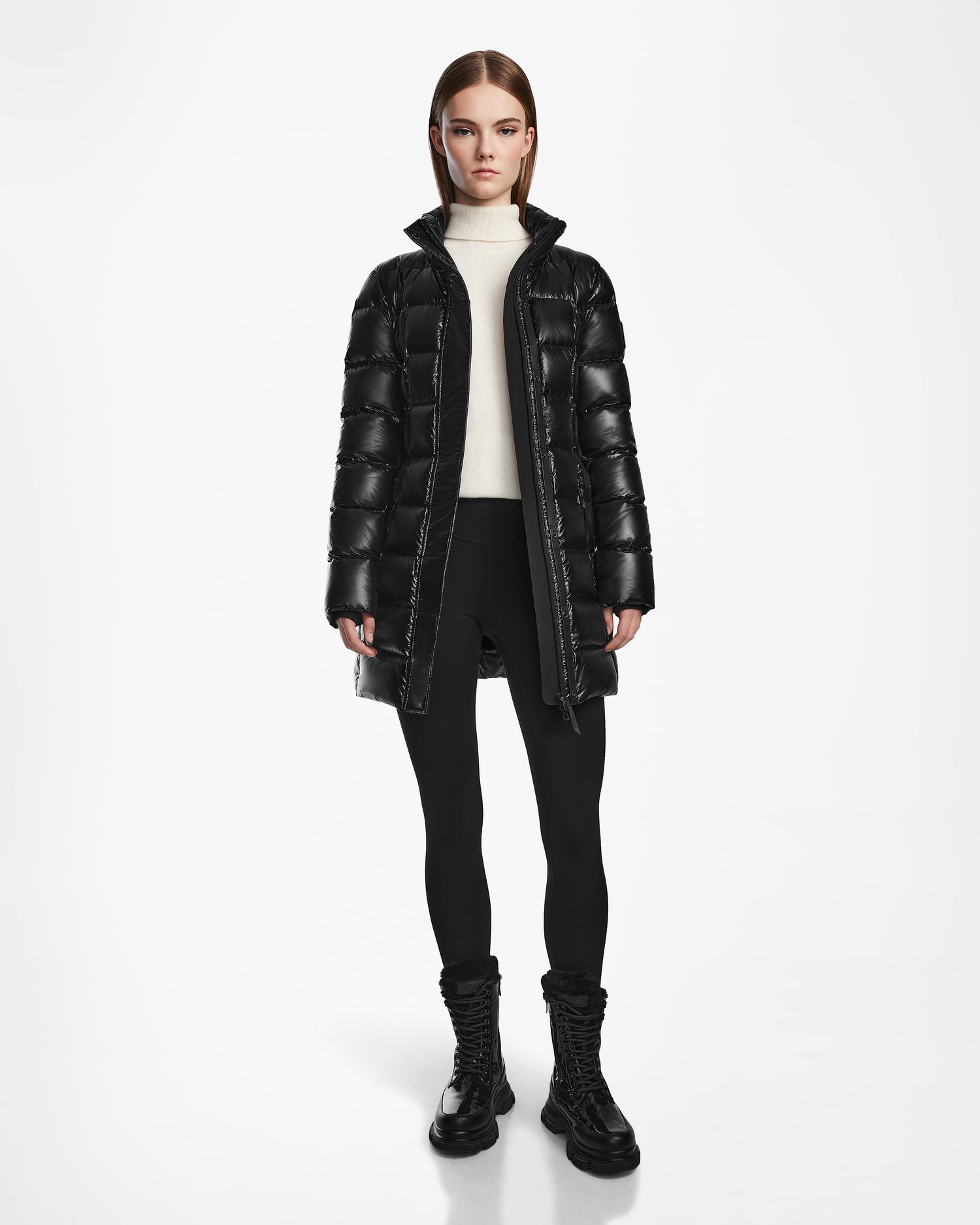 A woman with straight brown hair wears the Rudsak FRIDA S - L124526 Black mid-length down jacket with a removable hood, paired with a white turtleneck, black leggings, and lace-up boots against a plain white background.