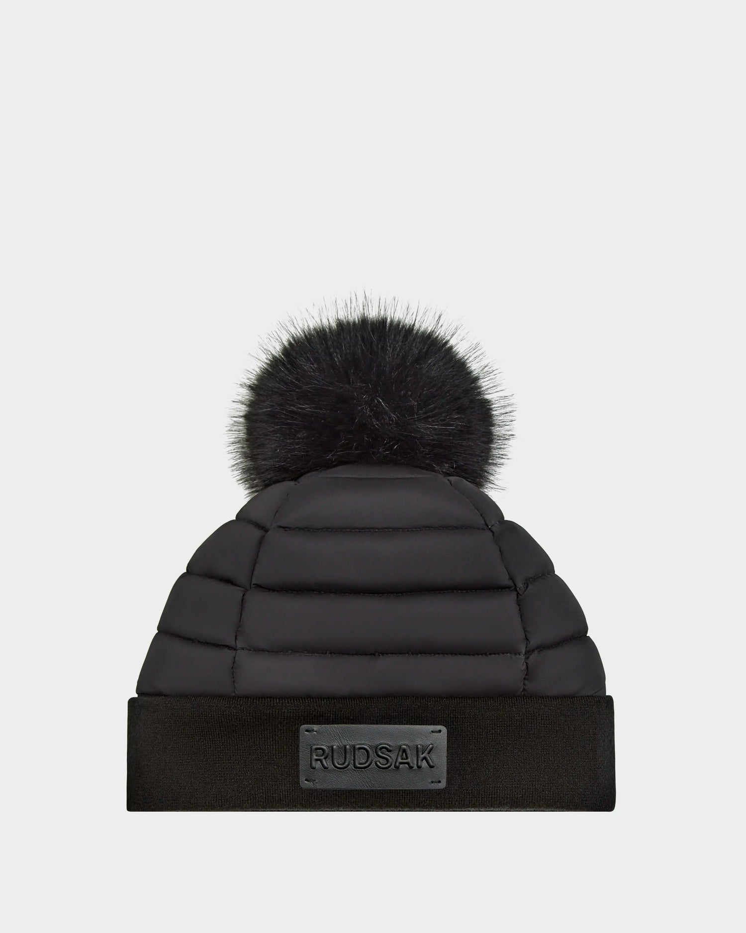 The GALA - 8424514 Black by Rudsak is a women’s quilted down puffer hat with a big faux fur pom pom and folded brim, finished with a front rectangular Rudsak patch—your go-to winter accessory.