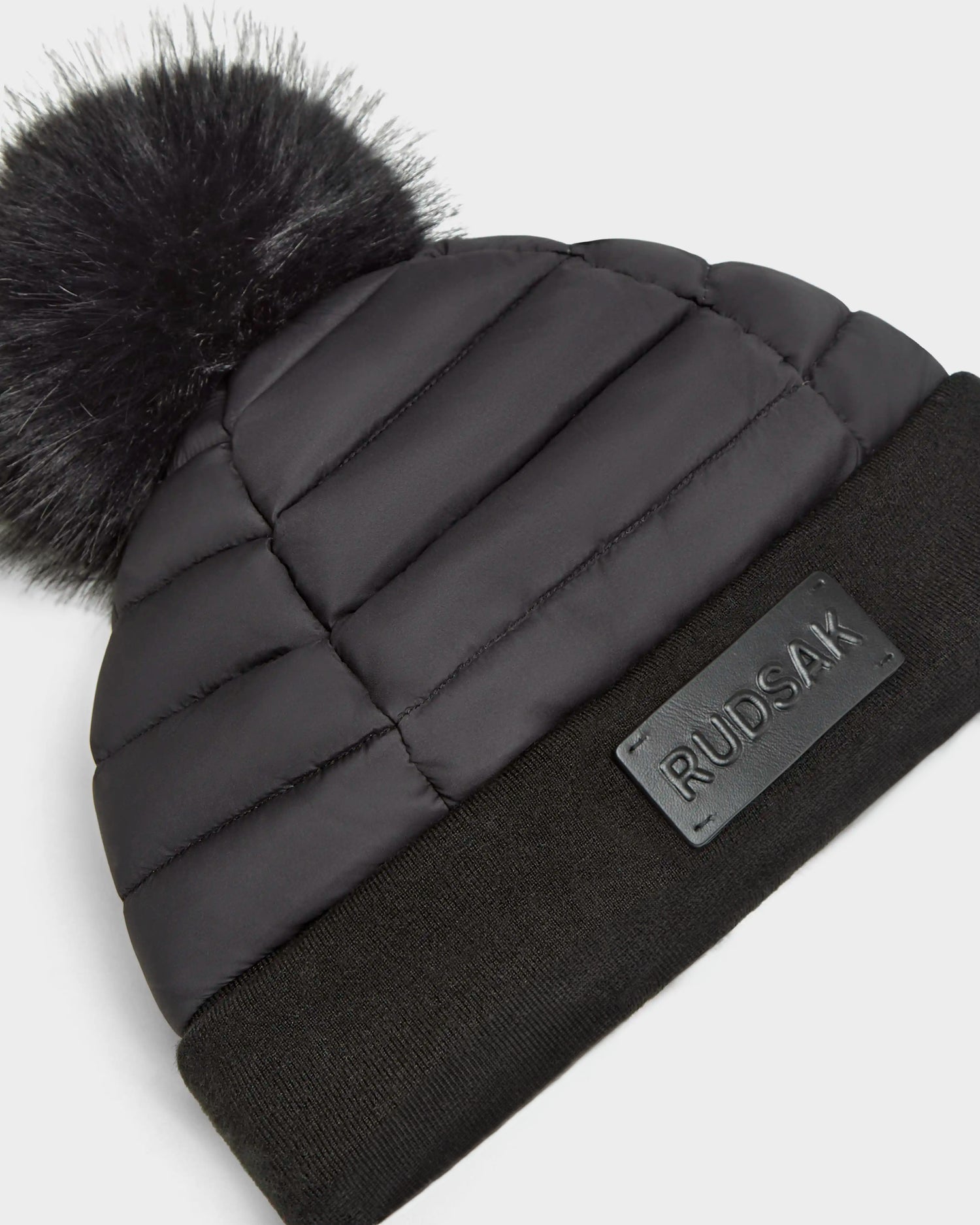 The GALA - 8424514 Black by Rudsak is a womens quilted down puffer hat with a wide ribbed cuff, large faux fur pom pom, and front Rudsak logo patch—a stylish go-to for winter warmth.