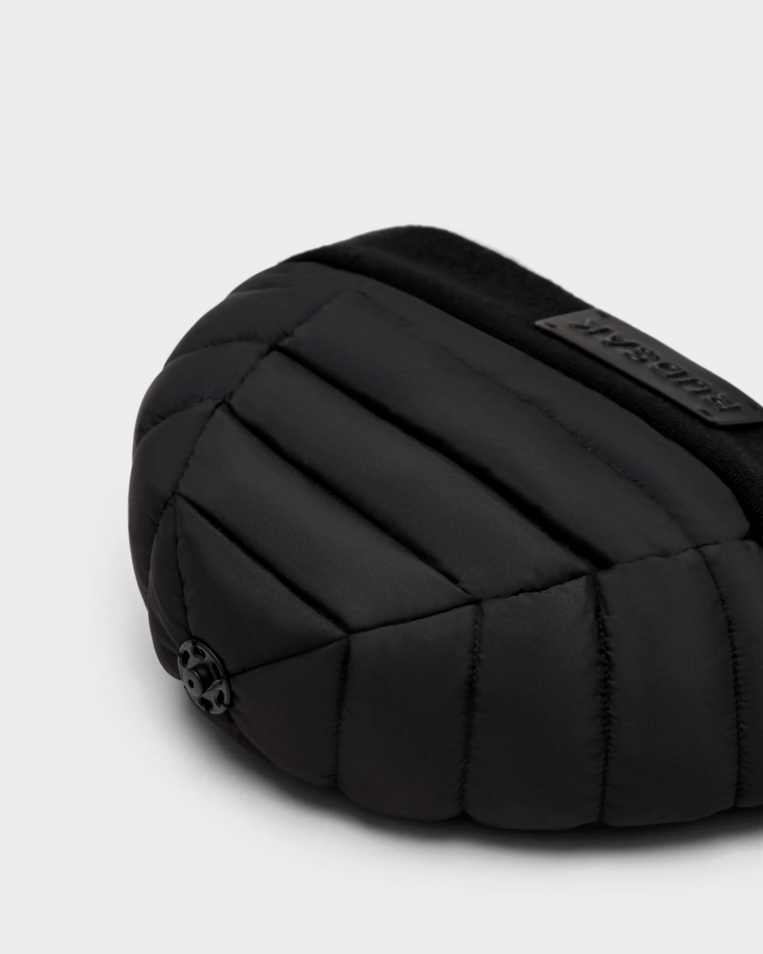 Close-up of Rudsaks GALA - 8424514 Black bag, featuring a quilted, padded design with a small round logo and a rectangular raised label, styled like a womens down puffer hat, set against a light gray background.