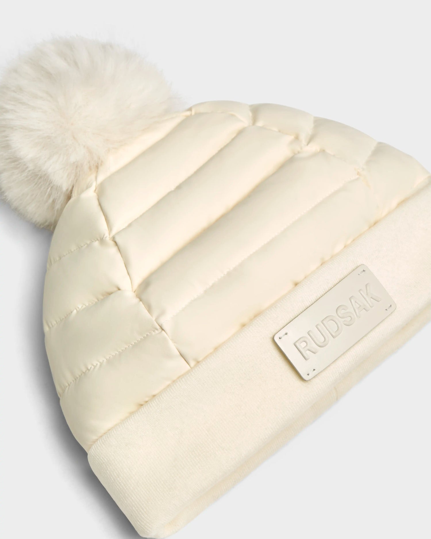 The GALA - 8424514 Iceland by Rudsak is a cream womens quilted down puffer hat with a wide knit brim, large faux fur pom pom, and a front rectangular RUDSAK patch—perfect for winter.