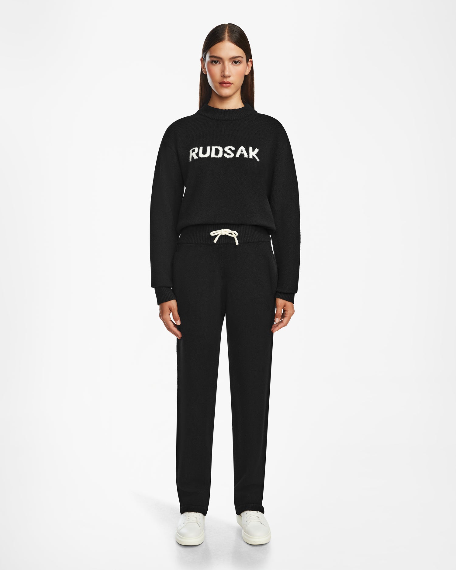 A woman with straight brown hair faces forward, wearing the Rudsak GEIGER - 9524512 Black W sweater with white RUDSAK lettering, relaxed lounge pants with a white drawstring, and white sneakers against a plain white background.