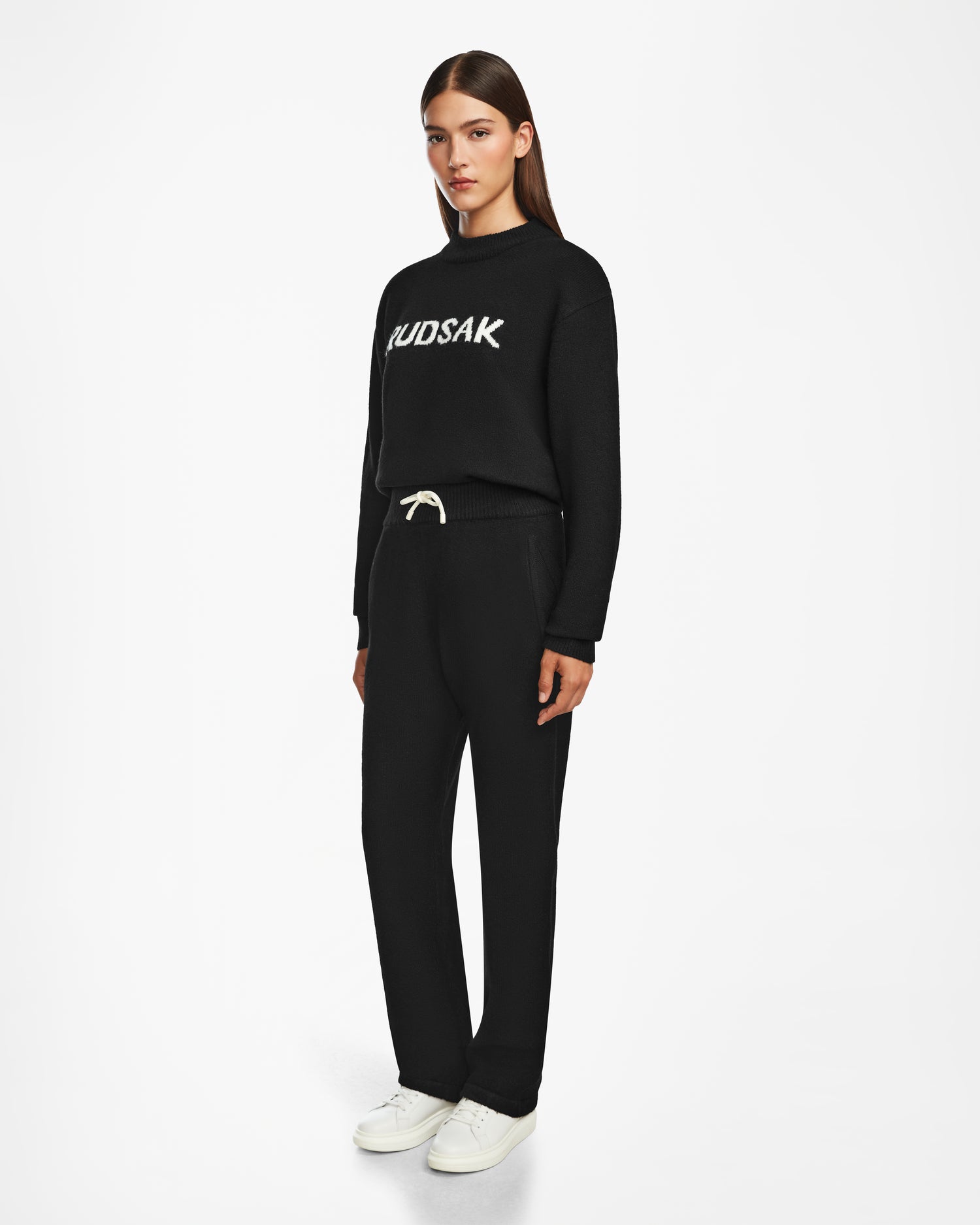 A woman with straight brown hair and a neutral expression wears the Rudsak GEIGER - 9524512 Black W pants with a white drawstring, a black MUDSAK sweater, and white sneakers against a white background.