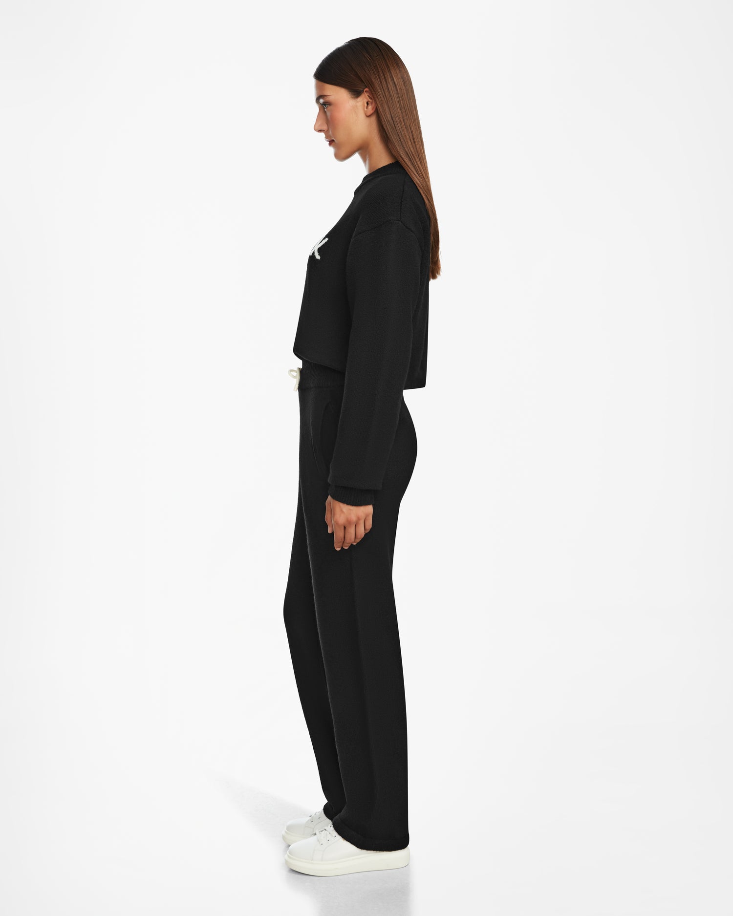 A woman in profile wears the Rudsak GEIGER - 9524512 Black W long-sleeve top with lounge pants and white sneakers, standing against a white background. She has straight long brown hair and a neutral expression.