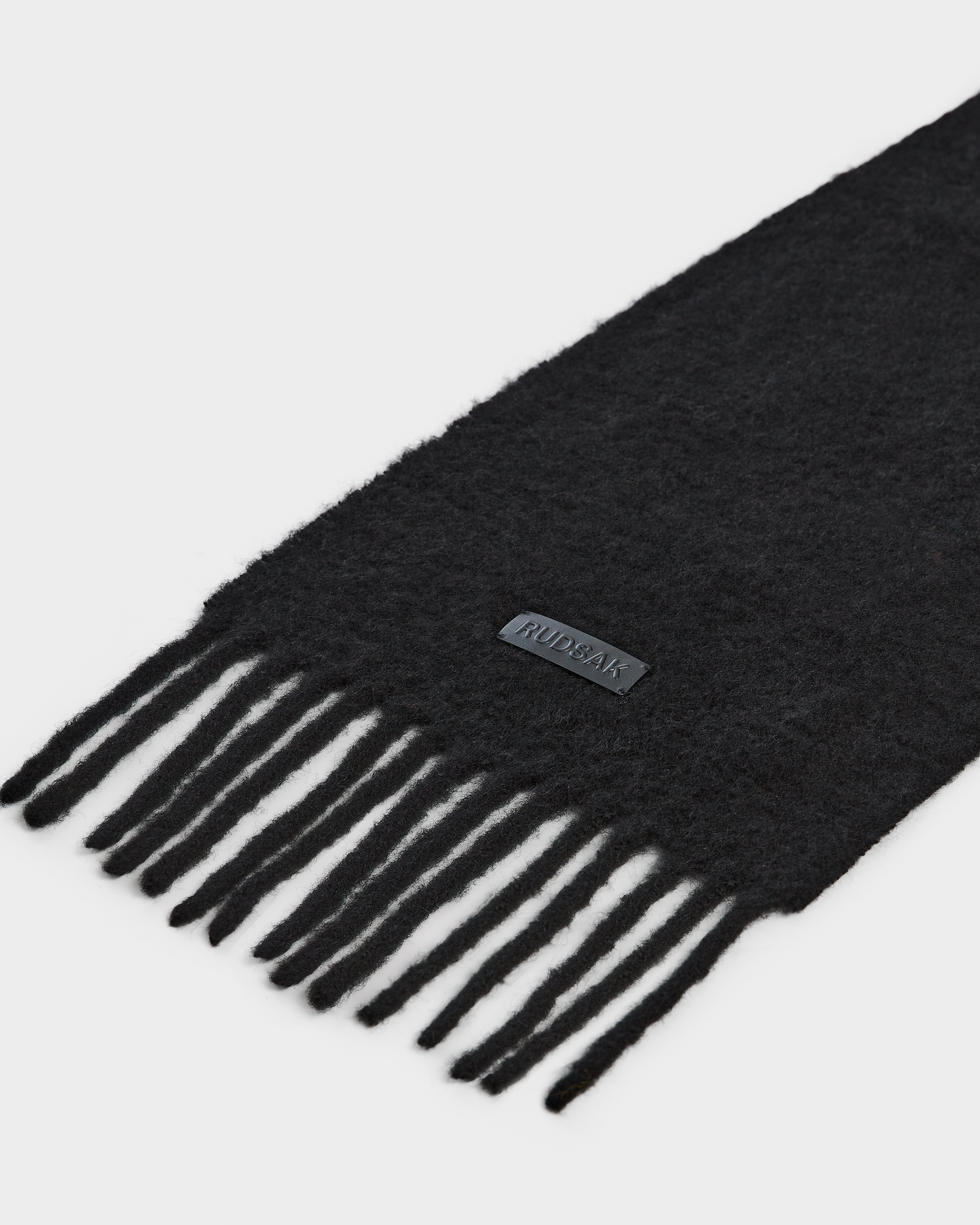 A close-up of the HAILEE - 9424507 Black scarf by Rudsak, crafted from black alpaca wool with a soft, fuzzy texture, fringed ends, and a small rectangular Rudsak label—an essential unisex winter accessory.