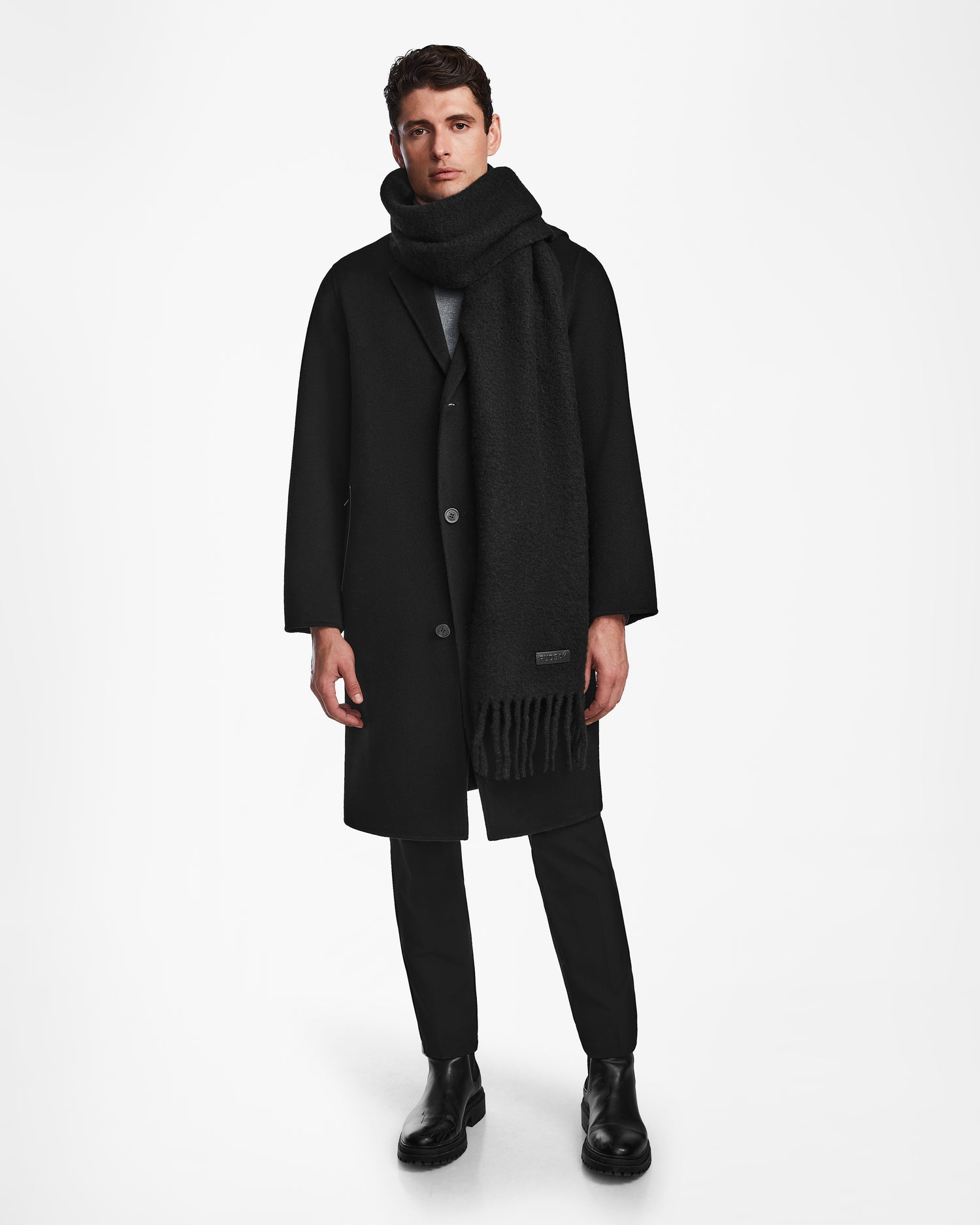 A man stands against a white background wearing the Rudsak HAILEE - 9424507 Black coat, black pants, and boots. An alpaca wool scarf wraps around his neck as he gazes forward with a neutral expression—a timeless winter look.