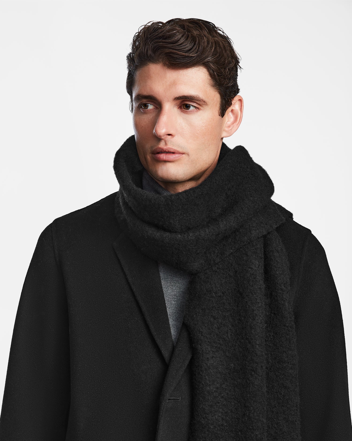 A man with short, wavy brown hair models the Rudsak HAILEE - 9424507 Black unisex scarf with a black coat, looking to the side against a plain white background and highlighting this cozy winter accessory.