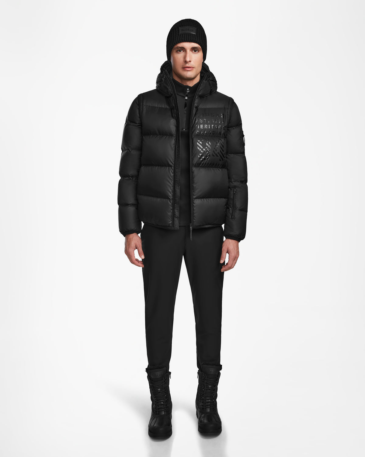 A man faces forward on a plain white background, wearing the Rudsak HANS - 6124518 Black mens down ski jacket with RECCO rescue tech, paired with black pants, boots, and a beanie.