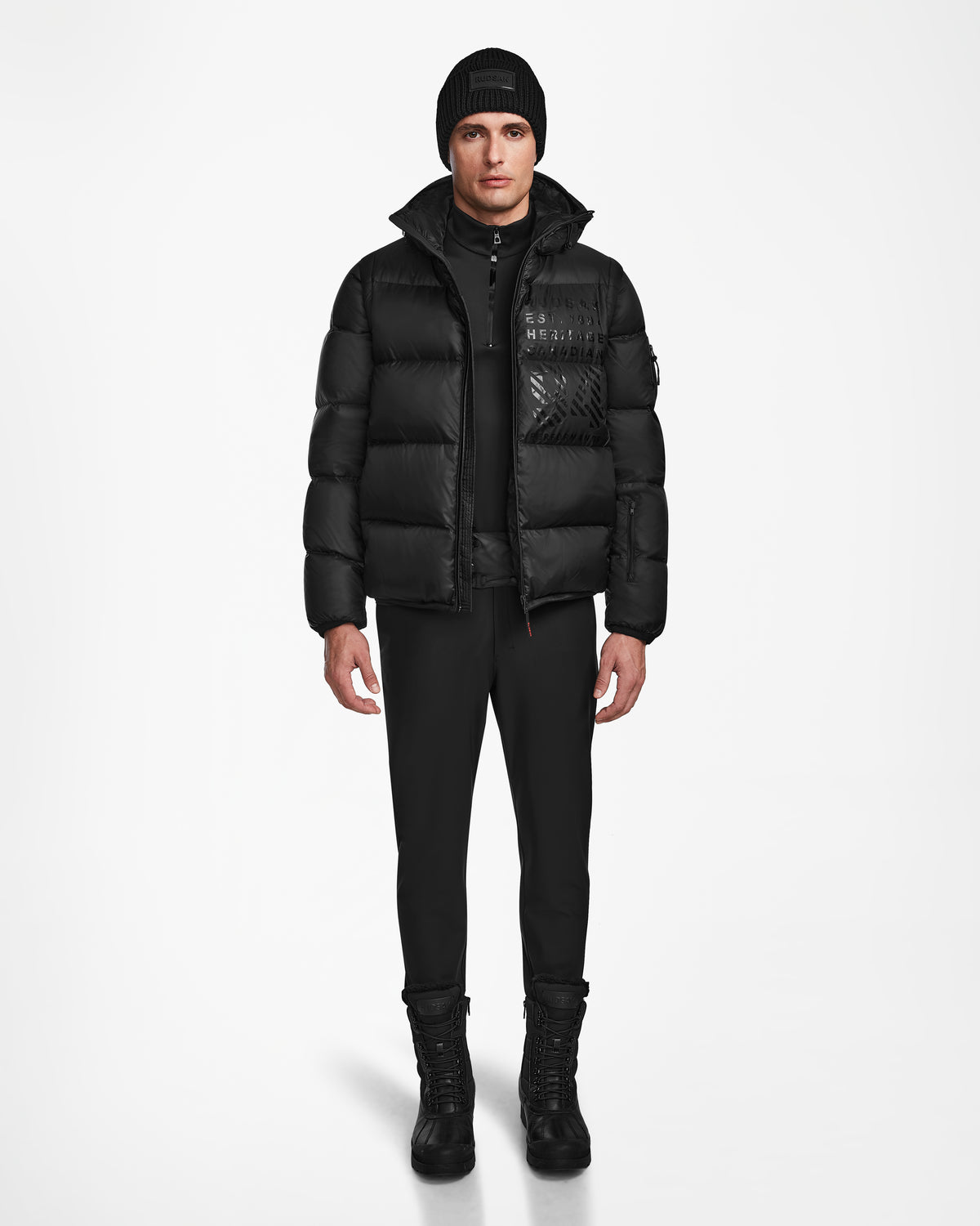 A man faces forward against a plain white background, dressed in the Rudsak HANS - 6124518 Black mens down ski jacket, paired with black pants, boots, and a knit beanie for winter protection.