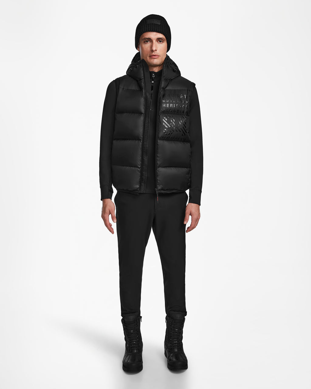 A man faces forward in Rudsak’s HANS - 6124518 Black: a black beanie and puffer vest with RECCO rescue tech, plus black zip-up jacket, pants, and boots—showcasing winter protection against a plain white background.