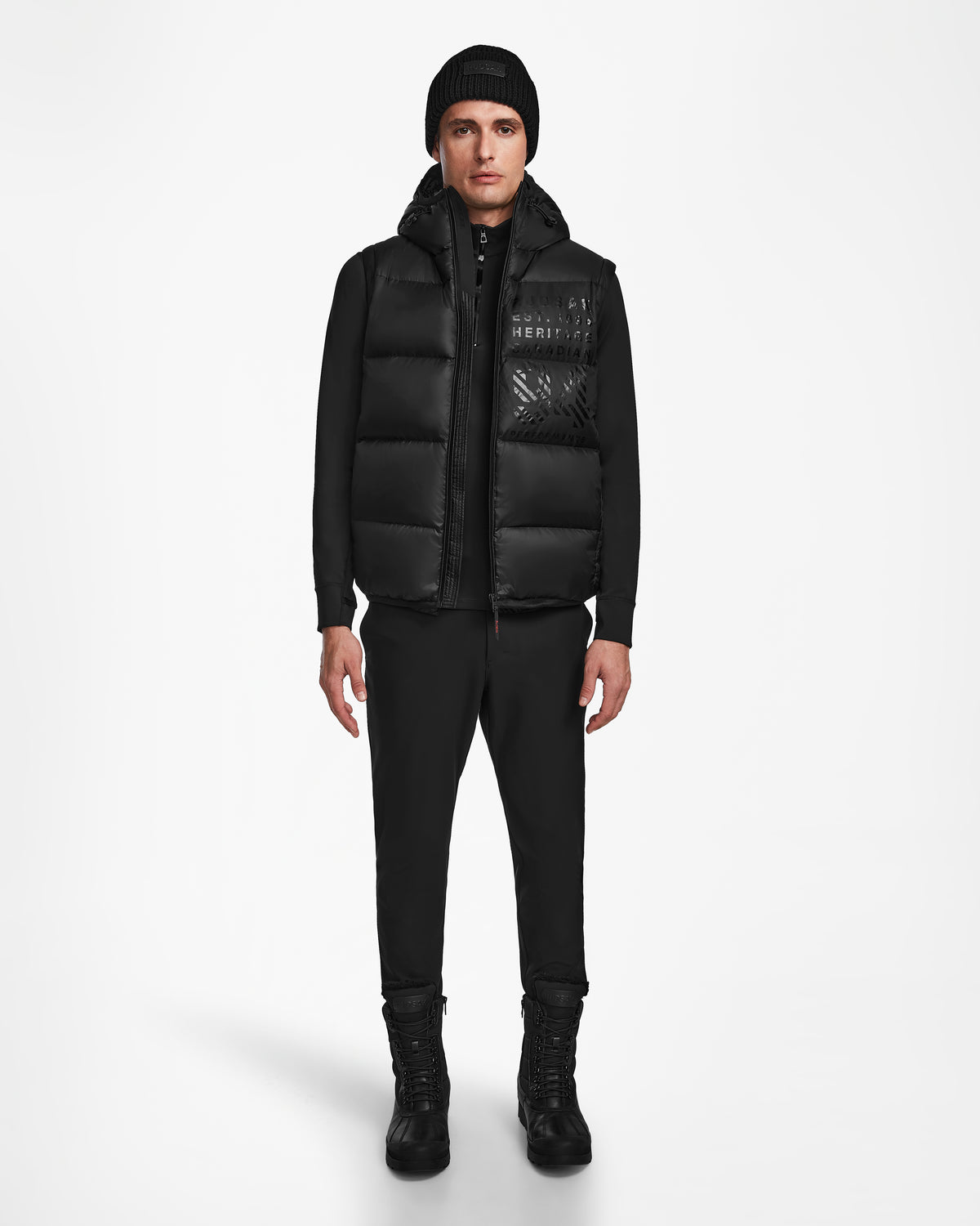 A man faces forward in Rudsak’s HANS - 6124518 Black: a black beanie and puffer vest with RECCO rescue tech, plus black zip-up jacket, pants, and boots—showcasing winter protection against a plain white background.