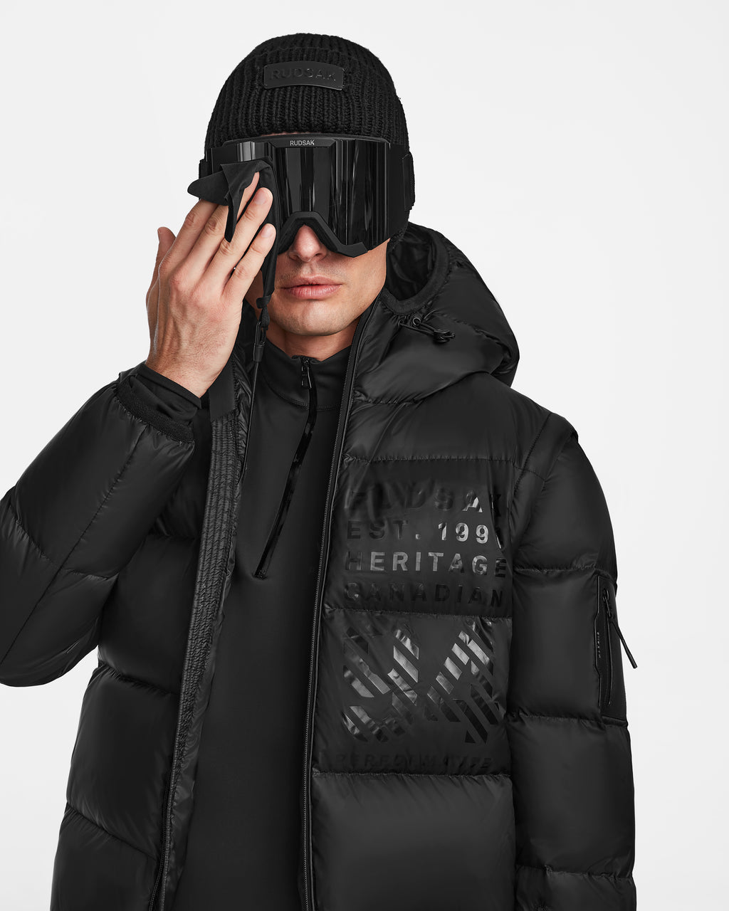 A person in a Rudsak HANS - 6124518 Black mens down ski jacket, black beanie, and large ski goggles cleans the goggles with a cloth against a plain white background.