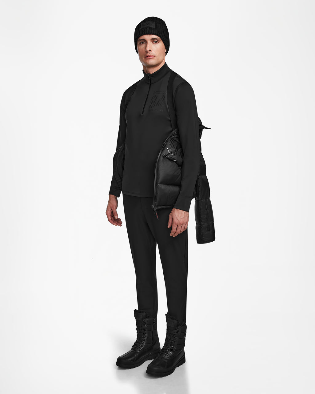 A person stands against a white background in all-black attire, holding the Rudsak HANS - 6124518 Black men’s down ski jacket with RECCO rescue technology over one shoulder.