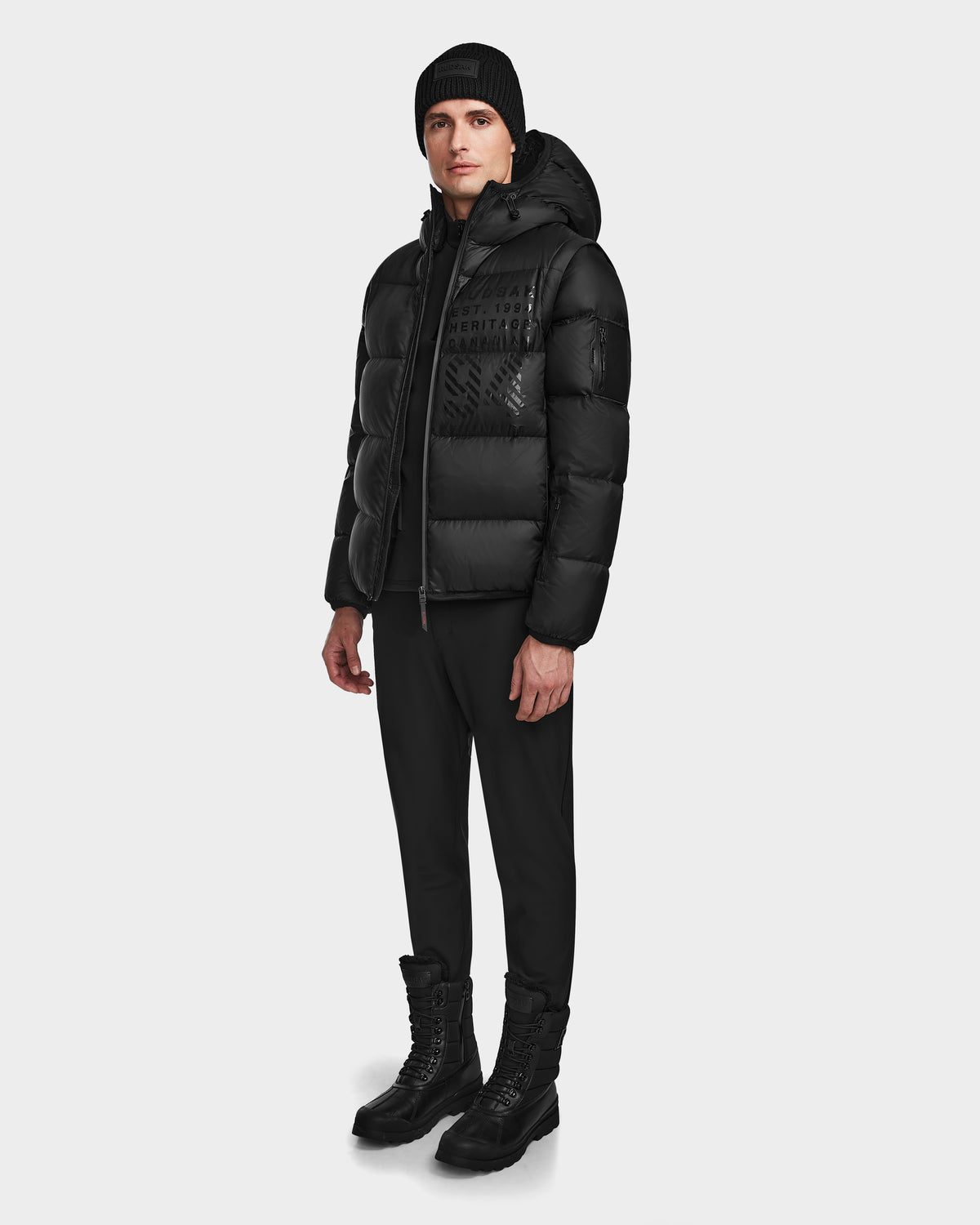 A man wearing the Rudsak HANS - 6124518 Black men’s down ski jacket with RECCO rescue technology, black pants, and black boots stands against a plain white background.