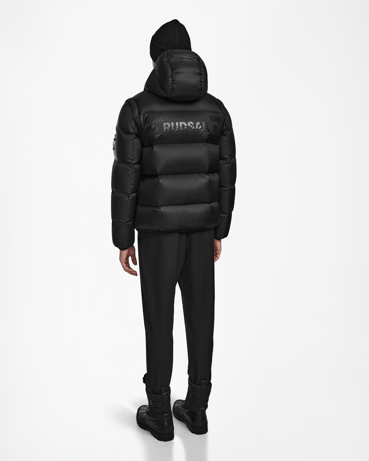 A person faces away, wearing the Rudsak HANS - 6124518 Black men’s down ski jacket with a design on the back, plus black pants, boots, and a knit beanie, all set against a plain white background.