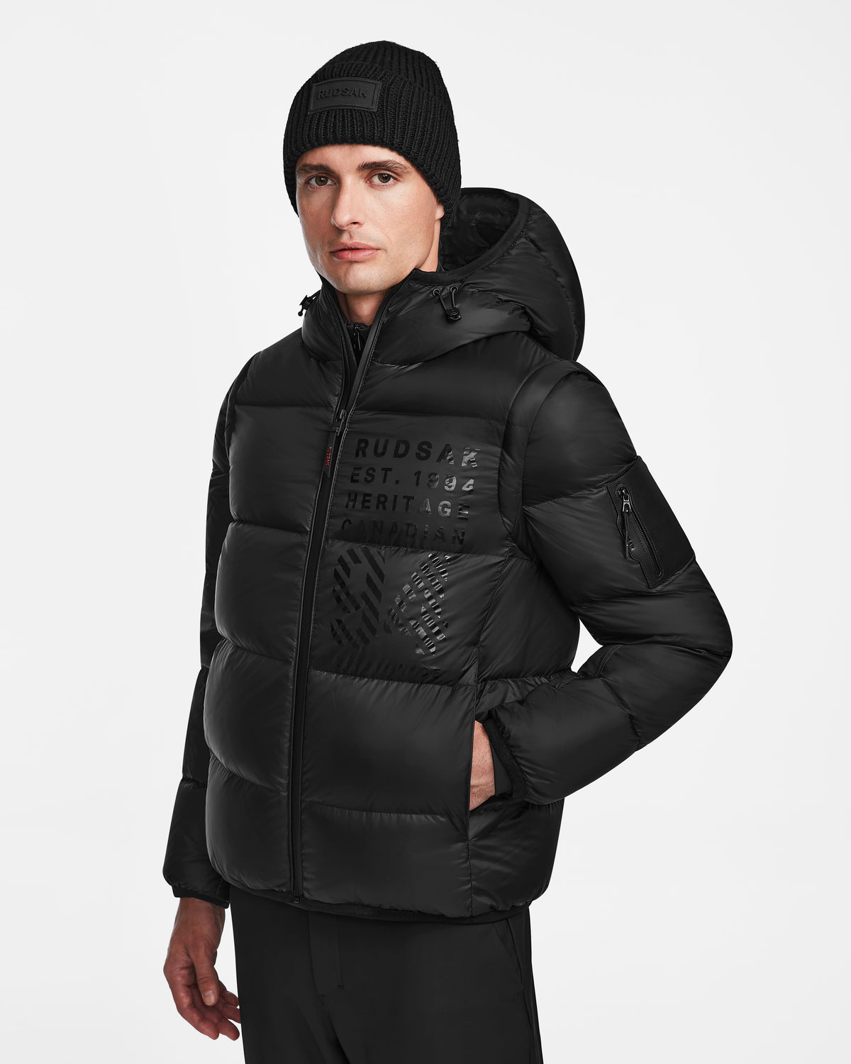 A man wears the Rudsak HANS - 6124518 Black men’s down ski jacket with wind and water resistance, RECCO rescue tech, logo/text details on the chest, a black knit beanie, and black pants against a plain white background.