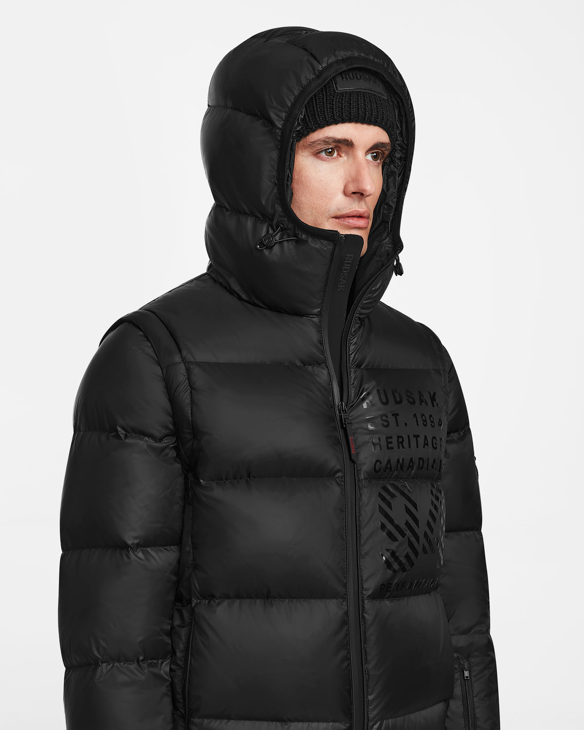 A person wears the Rudsak HANS - 6124518 Black mens down ski jacket with hood up and a black knit beanie, facing slightly right against a plain white background for optimal winter protection.