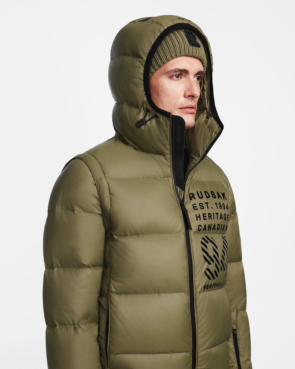 A man in the Rudsak HANS - 6124518 Loden Green men’s down ski jacket with a hood and matching knit beanie looks to the side. The wind- and water-resistant jacket features logo details, black text, and RECCO rescue technology.