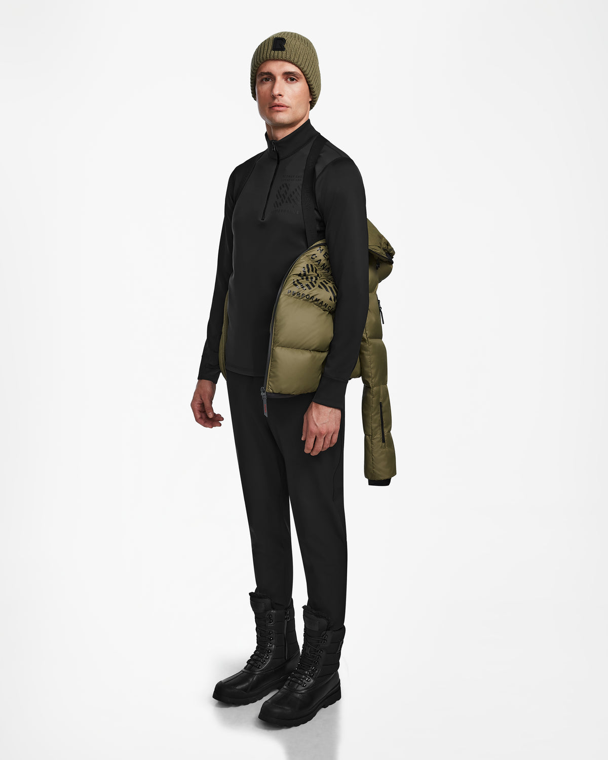 A man faces forward against a plain white background, wearing a black long-sleeve top, black pants, black boots, an olive green beanie, and the Rudsak HANS - 6124518 Loden Green wind and water-resistant jacket draped over one shoulder.