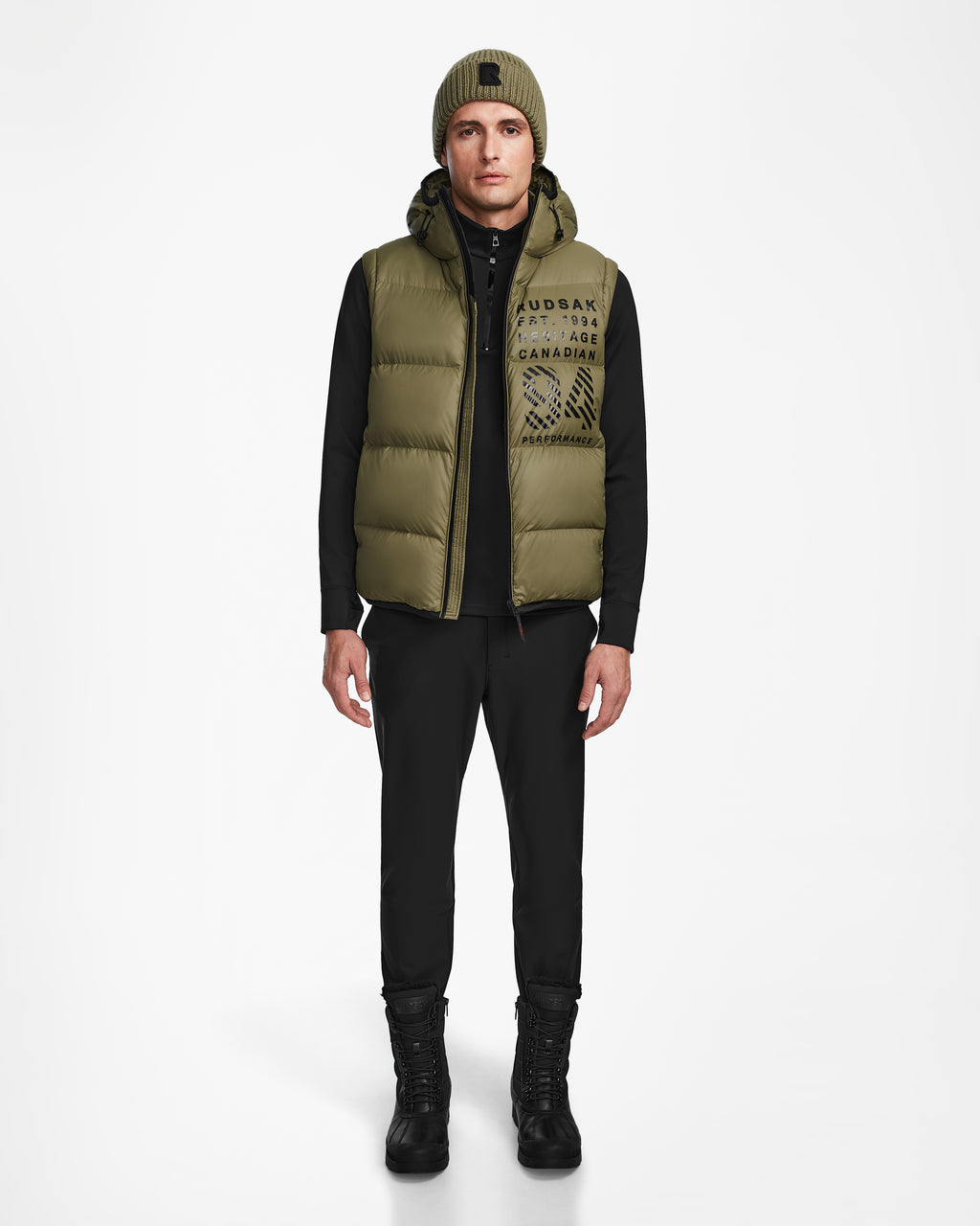 A man faces forward wearing the Rudsak HANS - 6124518 Loden Green wind and water-resistant puffer vest, styled with a green beanie, black long-sleeve shirt, black pants, and black boots, set against a plain white background.
