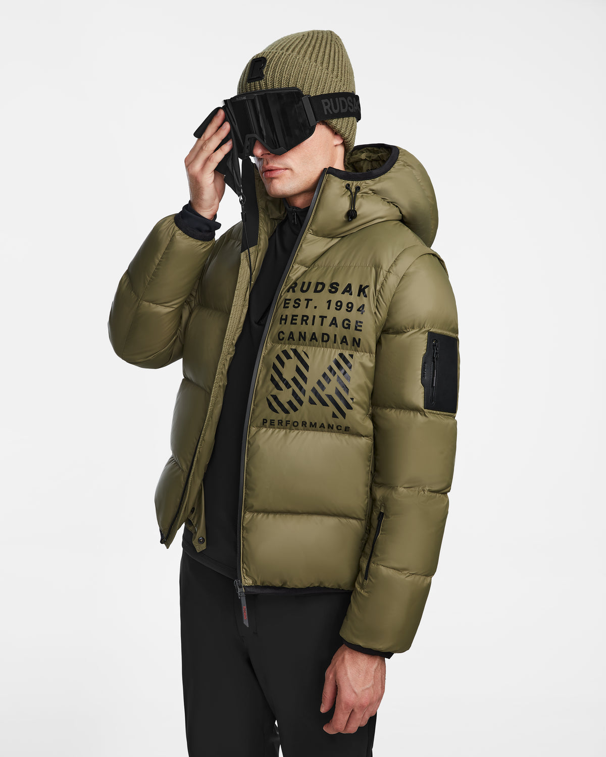 A person in the Rudsak HANS - 6124518 Loden Green men’s down ski jacket with RECCO rescue tech, olive hood, black pants, green beanie, and large ski goggles adjusts their goggles. The wind- and water-resistant jacket displays bold front graphics.