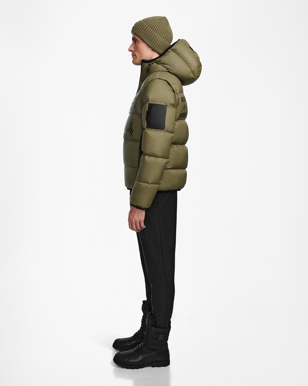 A man in profile against a white background wears the Rudsak HANS - 6124518 Loden Green wind- and water-resistant puffer jacket with a hood, paired with black pants, black boots, and a green knit beanie.