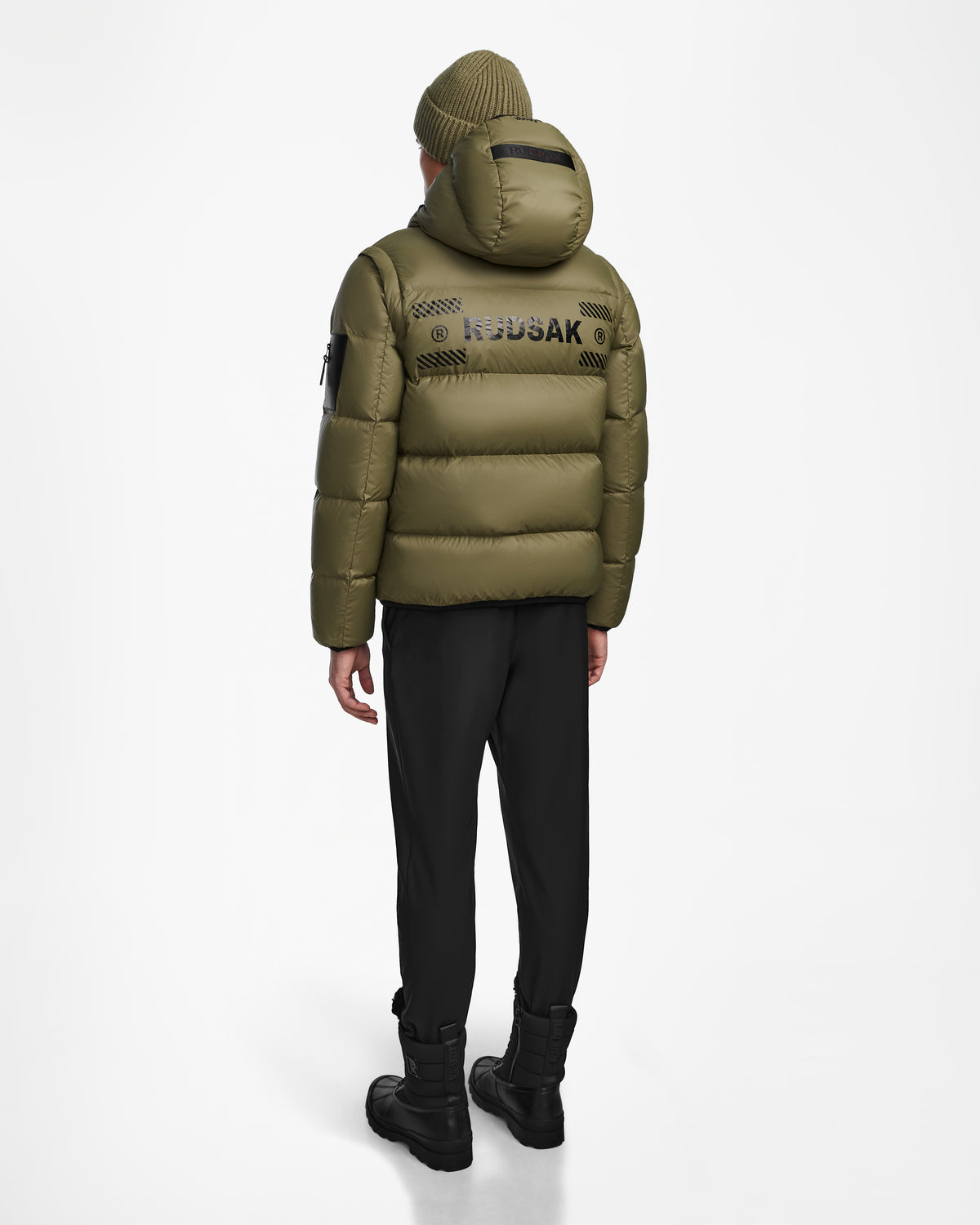 A person stands facing away, wearing Rudsak’s HANS - 6124518 Loden Green men’s down ski jacket with hood, black pants, boots, an olive knit beanie, and REDSKAP printed on the back of the wind- and water-resistant jacket.