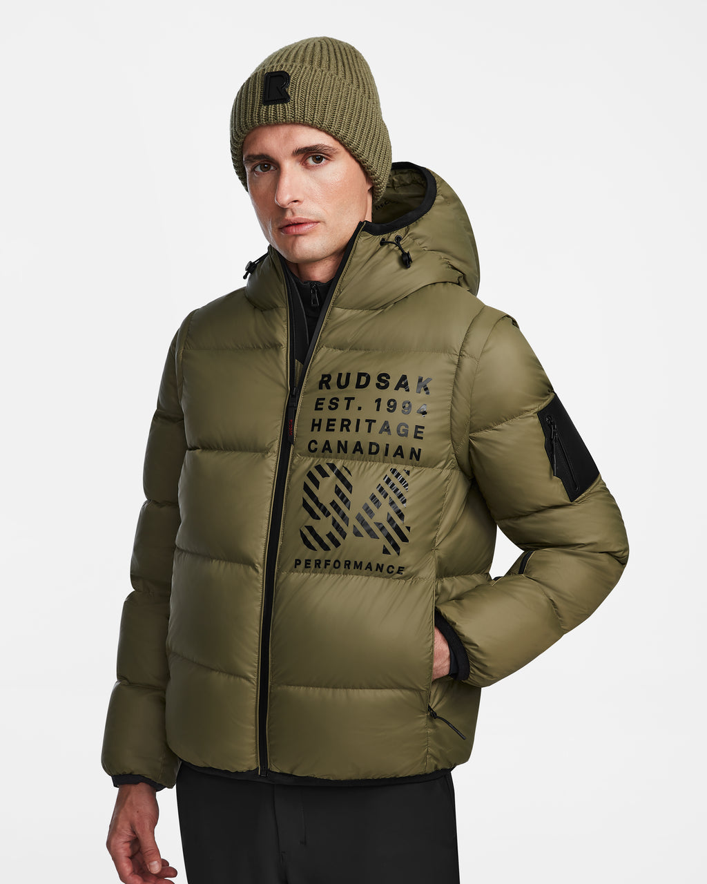 A man wears the Rudsak HANS - 6124518 Loden Green down ski jacket and matching knit beanie against a white background. The jacket features black lettering, patch details, and is wind and water-resistant for extra protection.