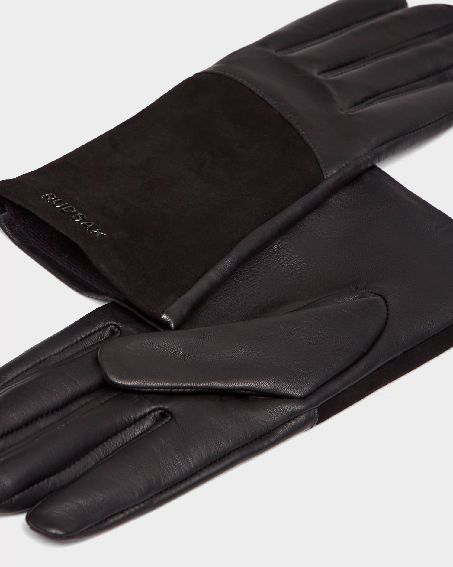 A pair of Rudsak HELENA - 8424529 Black womens leather gloves with a suede panel, wool lining, and embossed brand name on the cuff, shown on a light background.
