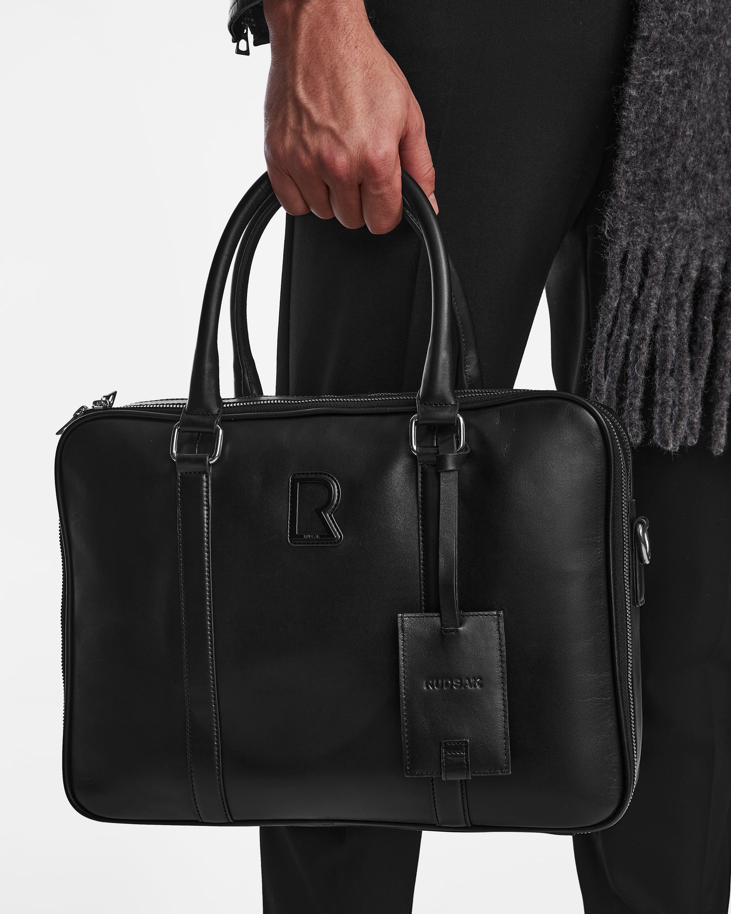 A person dressed in black holds the Rudsak HENRY L - 9323501 Black, a sleek unisex leather briefcase with two handles, adjustable shoulder strap, luggage tag labeled RUDSAK, and an embossed R logo on the front. Plain white background.