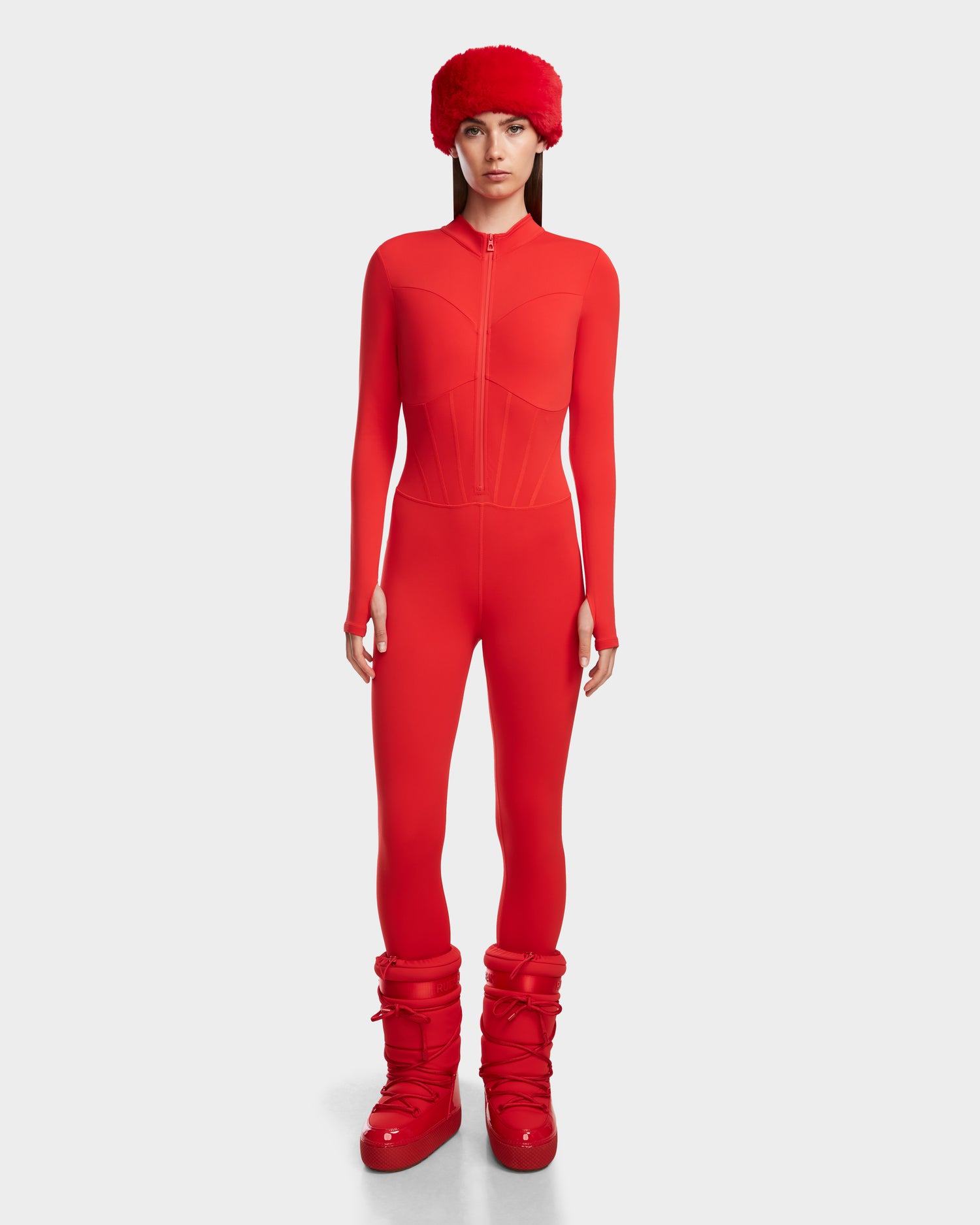 A person faces forward in the Rudsak IVY - 8524599 Flame Red womens stretch bodysuit, paired with a matching furry hat and red puffer boots, creating a bold ski-chic look against a plain white background.