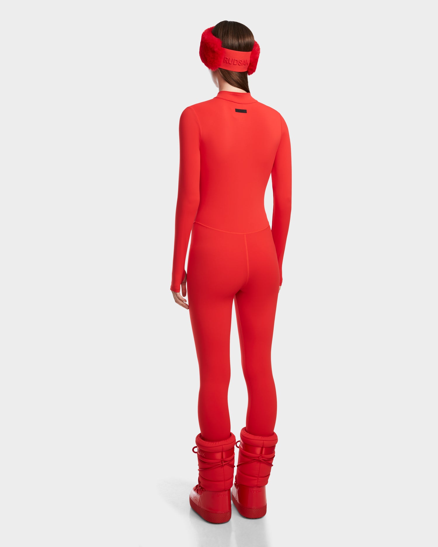 A woman faces away in a fitted, flame red IVY - 8524599 bodysuit by Rudsak, paired with matching earmuffs and boots. The plain white background emphasizes the sleek, ski-chic style of this modern monochrome look.