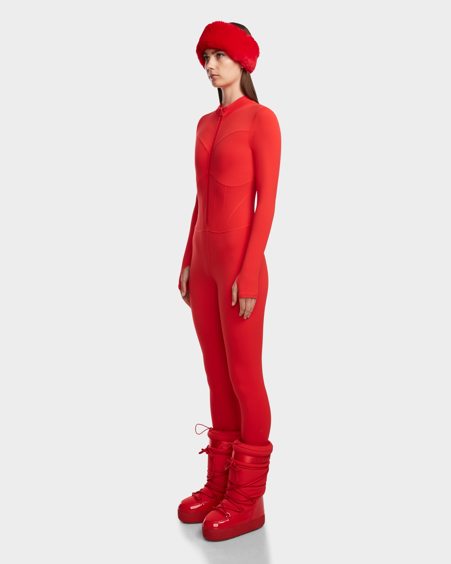 A woman stands in profile against a light gray background, wearing the Rudsak IVY - 8524599 bodysuit in Flame Red, paired with a red fuzzy headband and shiny red winter boots for a ski-chic look.